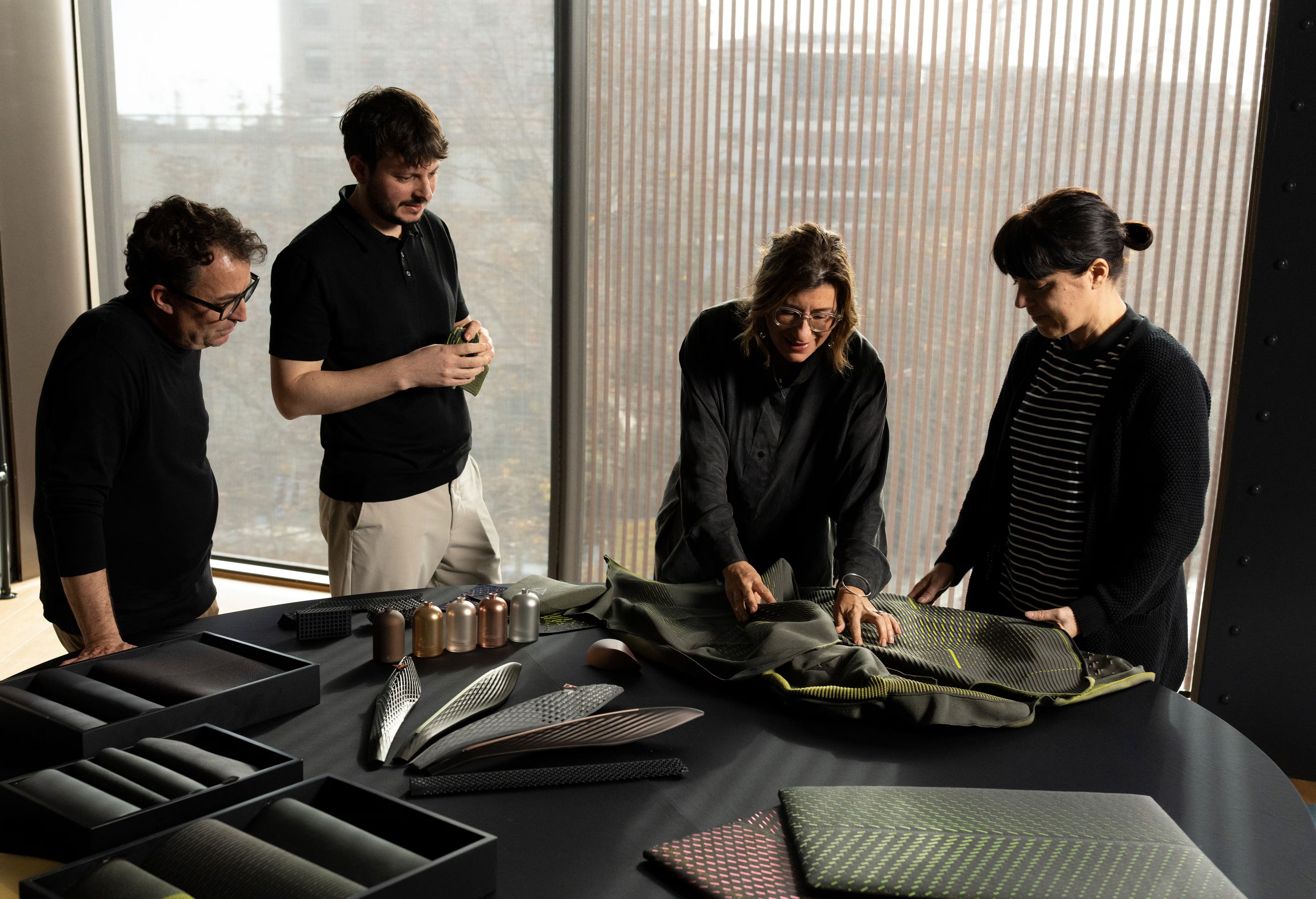 Štirje sodelavci stojijo ob mizi in pregledujejo različne materiale ter vzorce za oblikovanje. Na mizi so razporejeni tekstili, 3D-strukturirani elementi in barvni vzorci, ki nakazujejo razvoj novih rešitev.