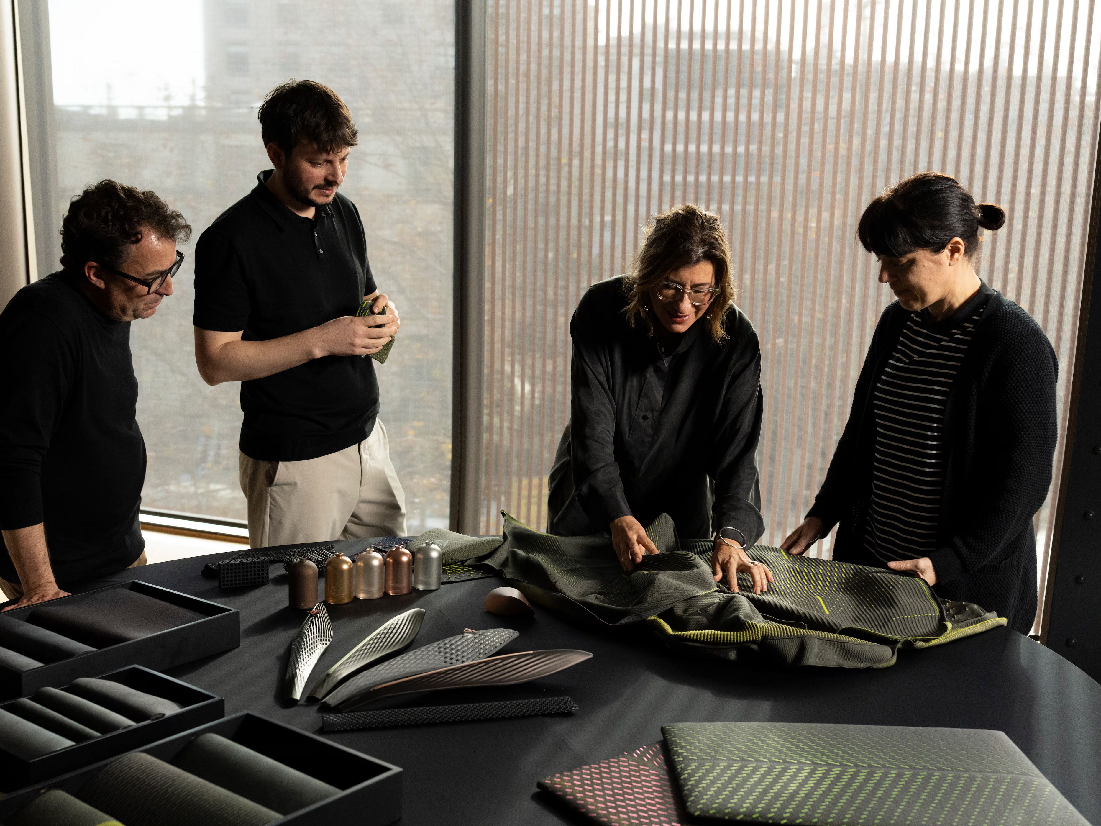 Štirje sodelavci stojijo ob mizi in pregledujejo različne materiale ter vzorce za oblikovanje. Na mizi so razporejeni tekstili, 3D-strukturirani elementi in barvni vzorci, ki nakazujejo razvoj novih rešitev.
