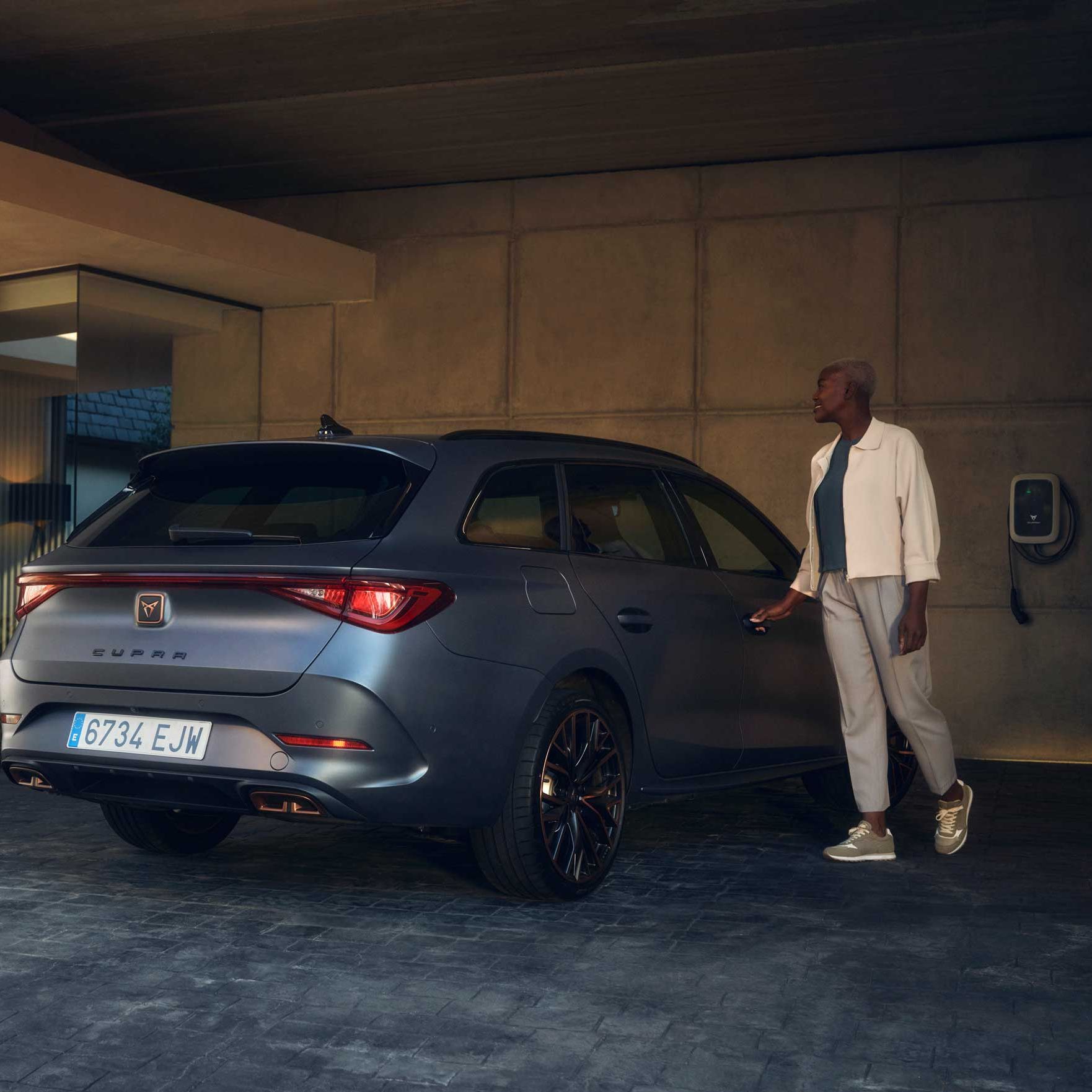 Ženska stoji ob sivem avtomobilu CUPRA, parkiranem pod modernim nadstreškom ob hiši. Drži v roki ključ in se približuje vozilu, v ozadju pa je na steni nameščena polnilna postaja. 