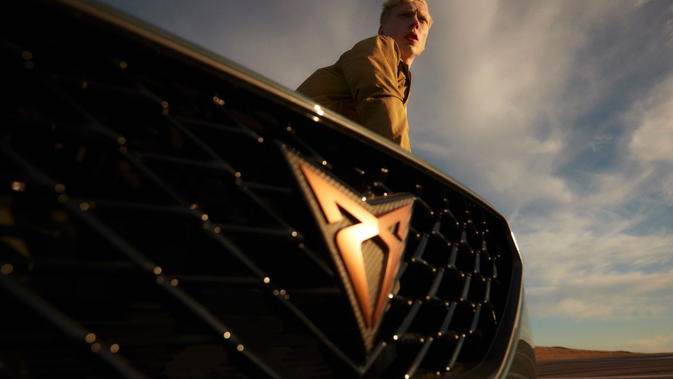 Moški stoji za avtomobilom CUPRA Formentor na katerem se jasno vidi logo znamke. Ozadje prikazuje modro-sive oblake na nebu.