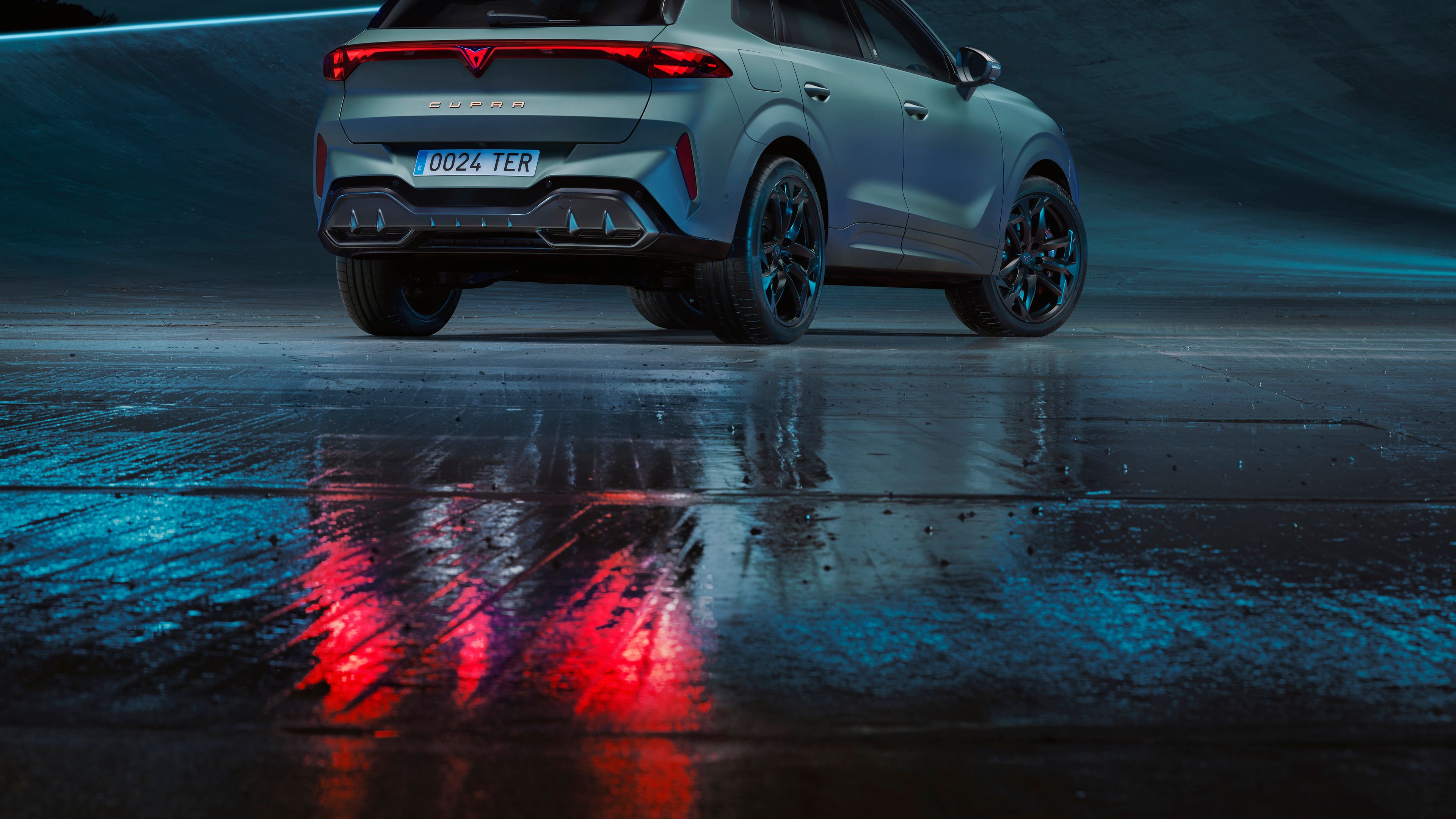 Nazaj kovinsko modrega mat avtomobila CUPRA Terramar parkiranega na mokri cesti. Luči avtomobila oddajajo rdečo svetlobo, ki se odbija na mokri površini cestišča. V ozadju je moderna arhitekturna struktura.