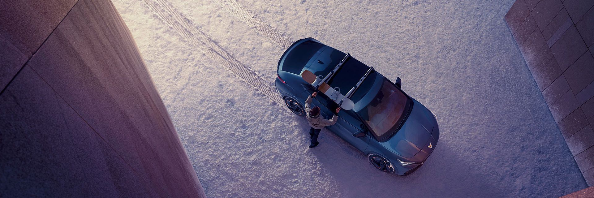 Blick von oben auf einen CUPRA Tavascan mit Snowboard Dachträger. Ein Mann montiert ein Snowboard darauf.