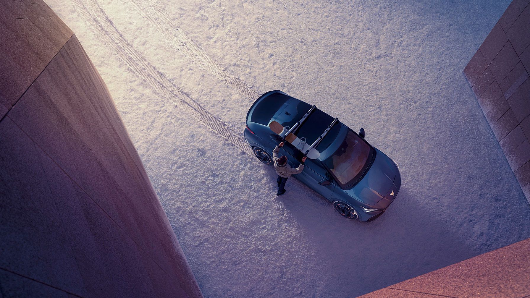 Pogled od zgoraj na osebo, ki stoji ob parkiranem avtomobilu CUPRA Tavascan na zasneženem tleh med dvema velikima zgradbama. Oseba namešča desko za snowboard na strešne nosilce vozila.