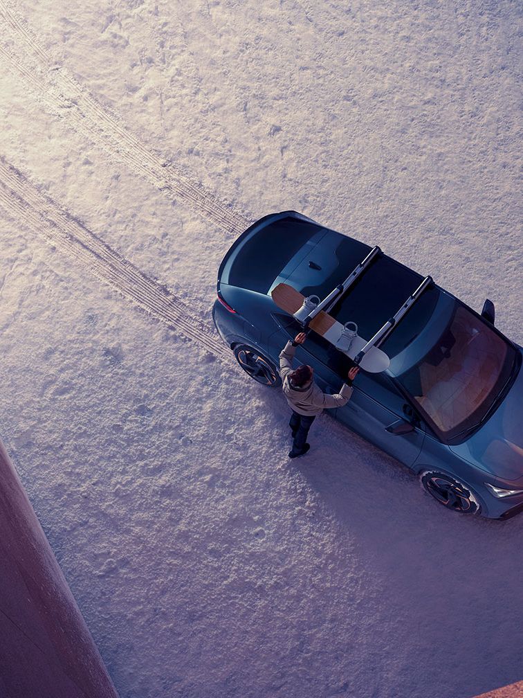 Pogled od zgoraj na osebo, ki stoji ob parkiranem avtomobilu CUPRA Tavascan na zasneženem tleh med dvema velikima zgradbama. Oseba namešča desko za snowboard na strešne nosilce vozila.