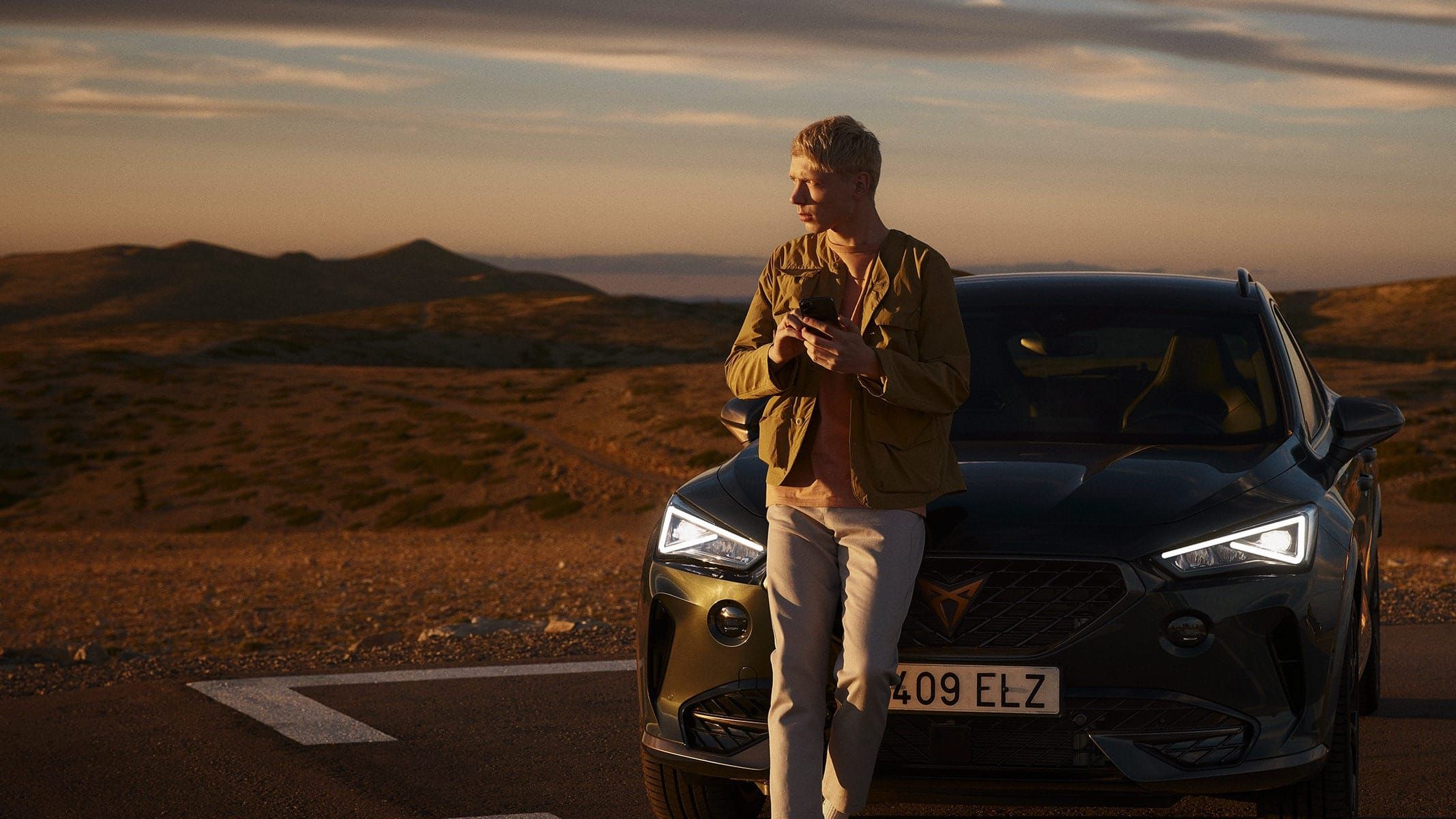 Moški stoji pred črnim avtomobilom CUPRA Umbrella Formentor na odprti cesti ob sončnem zahodu. Pokrajina je hribovita z zlatimi in zelenimi odtenki. 
