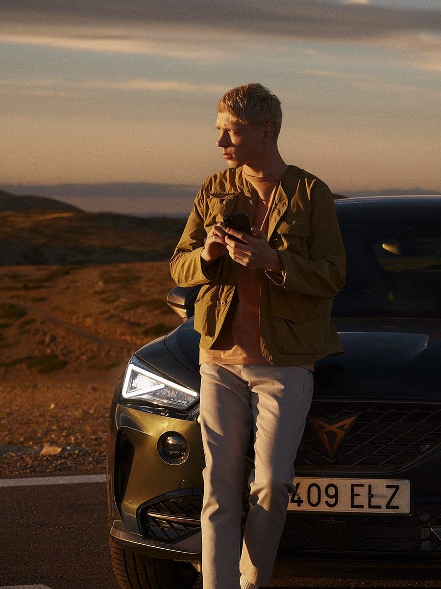 Moški stoji pred črnim avtomobilom CUPRA Umbrella Formentor na odprti cesti ob sončnem zahodu. Pokrajina je hribovita z zlatimi in zelenimi odtenki. 