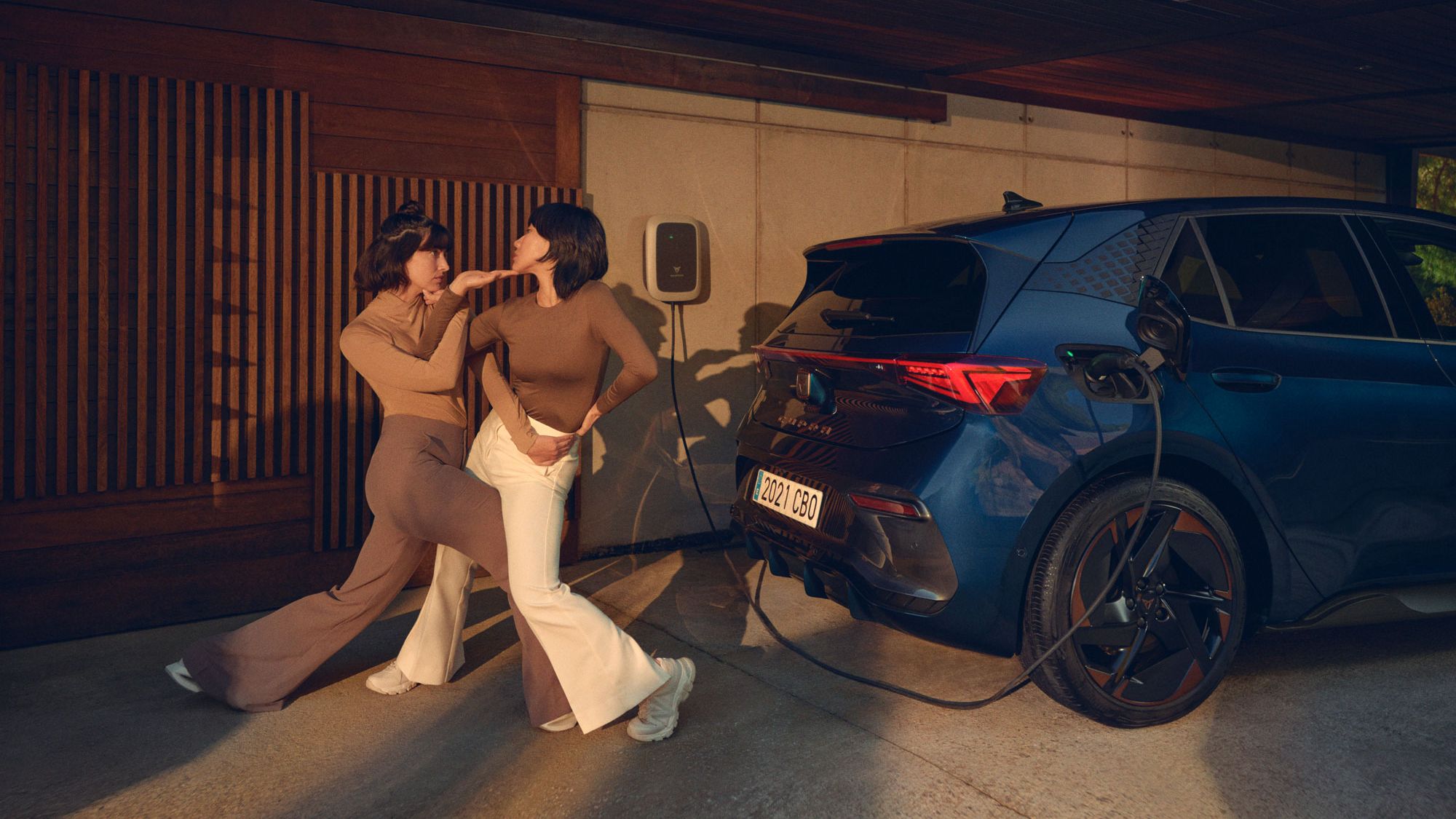 Dve osebi stojita v plesnem položaju ob parkiranem električnem avtomobilu, ki se polni na polnilni postaji. Vozilo je parkirano v garaži ob sodobni hiši.