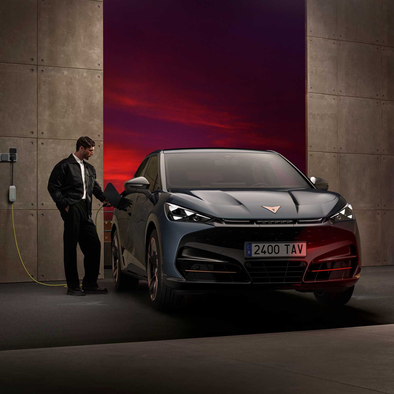 Simpatičen moški polni električni Cupra Tavascan v garaži ob sončnem zahodu. Avtomobil je priključen na polnilno postajo na steni. V ozadju so betonske stene in dramatičen rdeče-vijoličen večerni nebom.