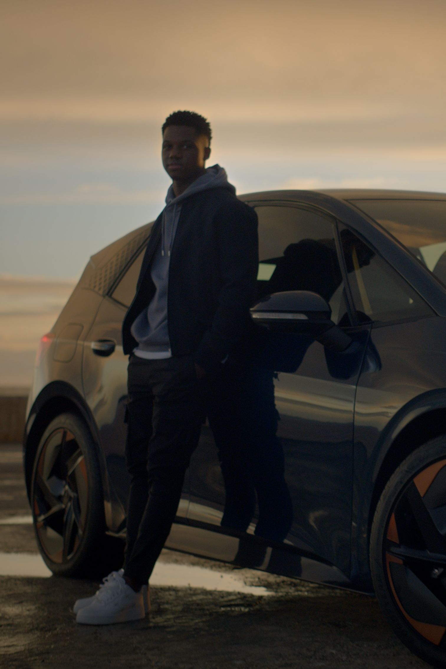 Nogometaš FC Barcelone Ansu Fati stoji naslonjen na CUPRA Borna ob oblačnem sončnem zahodu.