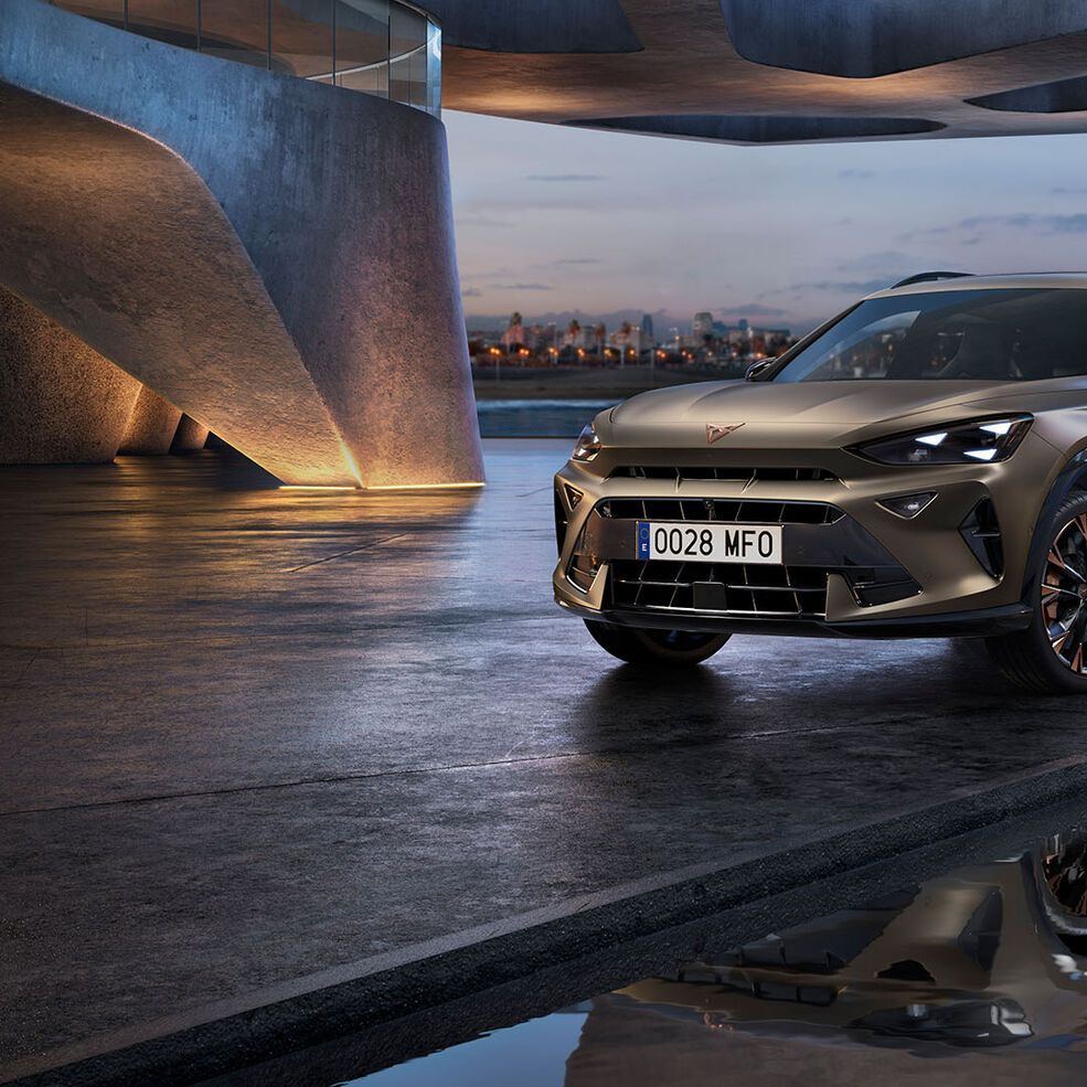 Moderen Cupra Formentor SUV je parkiran v urbanem okolju ob vodi, kjer se odraža v mirni vodi. Ozadje prikazuje mestne stavbe in visok stolp ob mestni obali v večernih urah.