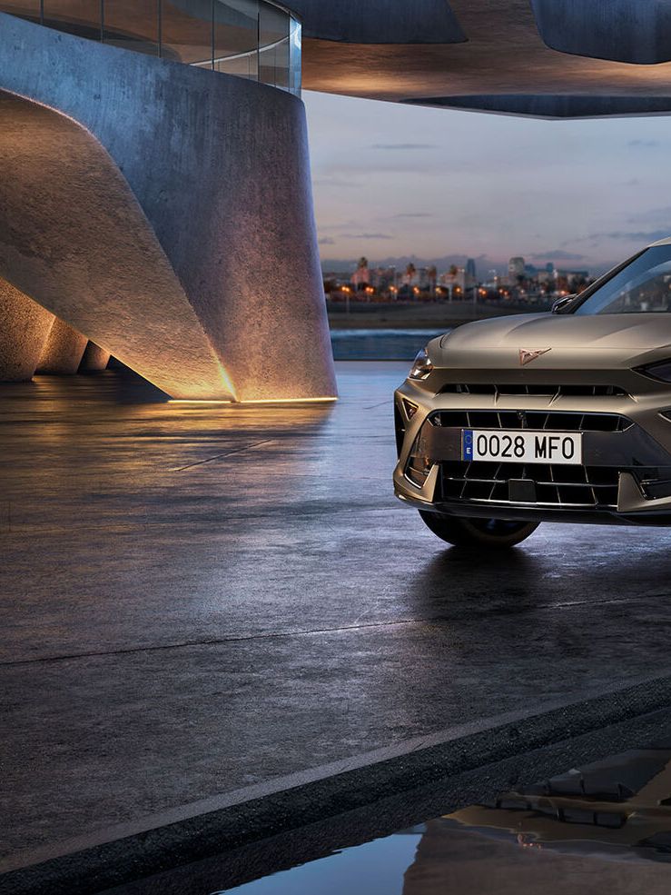 Moderen Cupra Formentor SUV je parkiran v urbanem okolju ob vodi, kjer se odraža v mirni vodi. Ozadje prikazuje mestne stavbe in visok stolp ob mestni obali v večernih urah.