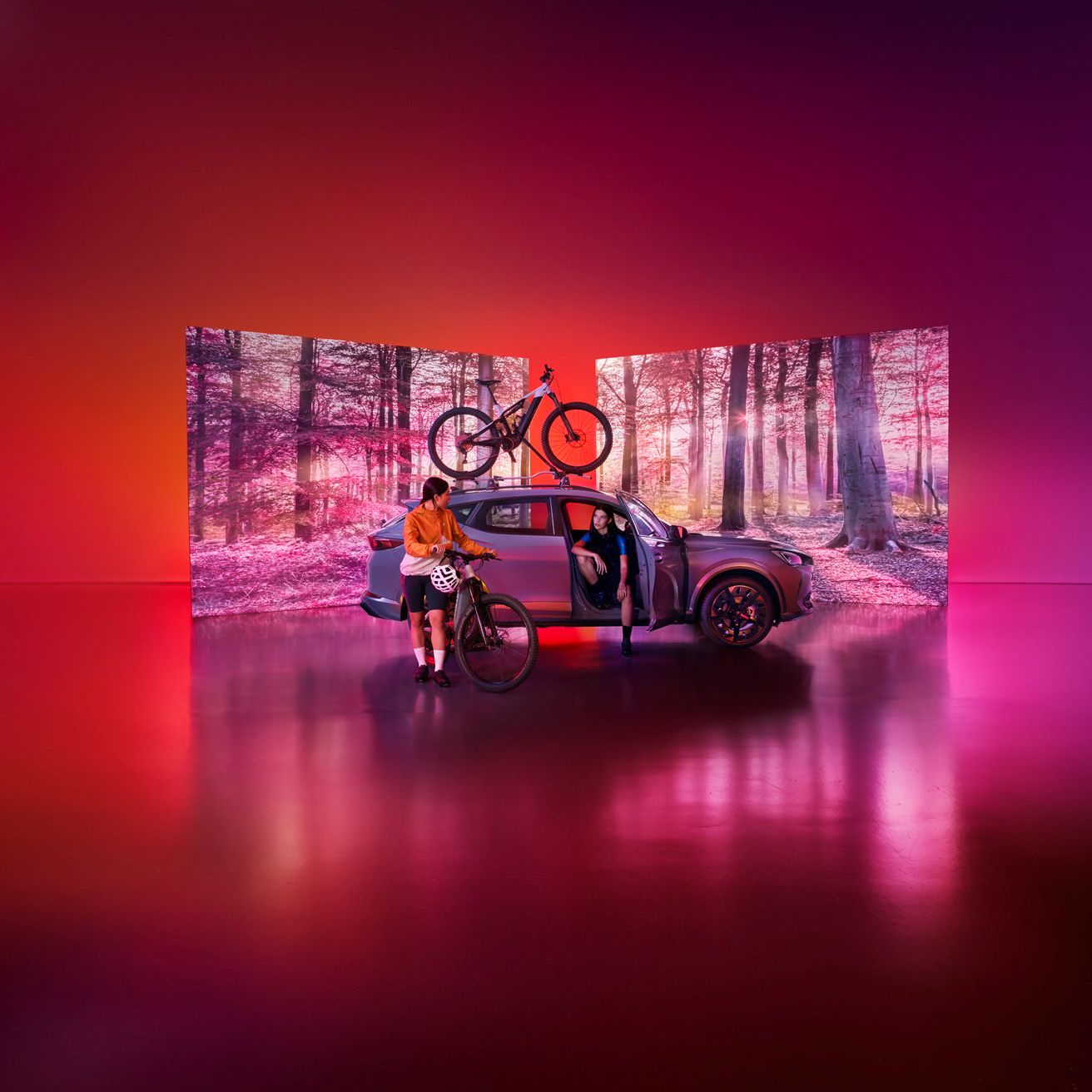 Ena ženska stoji ob avtomobilu CUPRA Formentor in drži kolo, druga sedi na sprednjem sedežu. Na strešnem nosilcu je nameščeno še eno kolo. Studio je osvetljen s toplimi rdečimi in rožnatimi toni, v ozadju pa so zasloni s podobo čudovitega gozda in toplim ambientnim svetlobnim ozračjem.