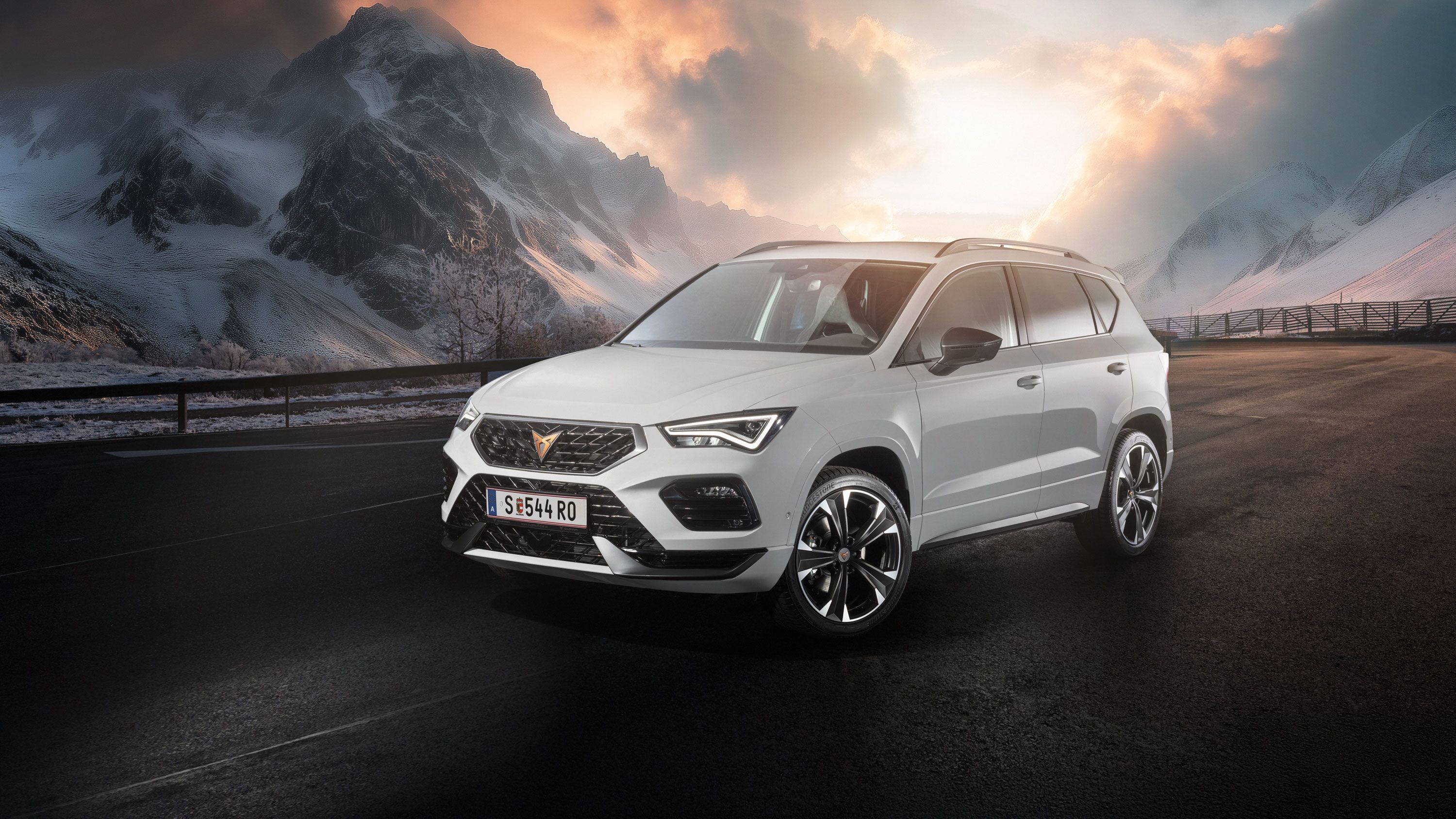 Ein weißer CUPRA Ateca fährt auf einer Bergstraße bei Sonnenaufgang. Die Straße ist nass und von Schnee gesäumt. Im Hintergrund sind schneebedeckte Berge und bewölkter Himmel zu sehen.