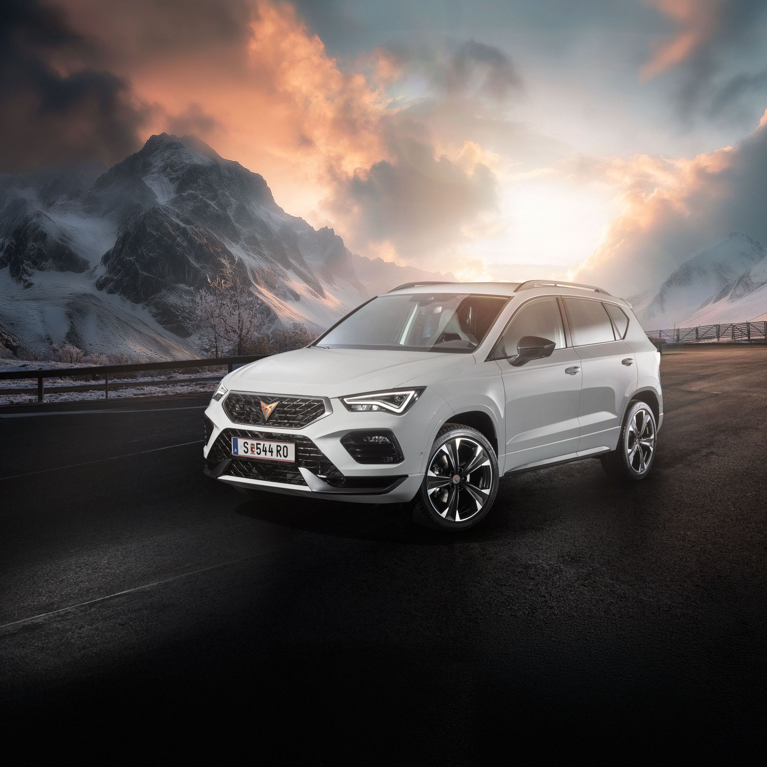 Ein weißer CUPRA Ateca fährt auf einer Bergstraße bei Sonnenaufgang. Die Straße ist nass und von Schnee gesäumt. Im Hintergrund sind schneebedeckte Berge und bewölkter Himmel zu sehen.