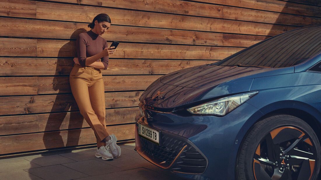 Ženska stoji ob leseni steni in drži mobilni telefon. Ob njej je parkiran moder CUPRA Born z oranžnimi platišči.