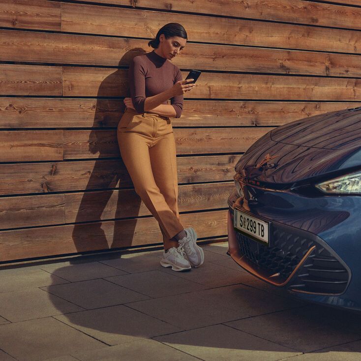 Ženska stoji ob leseni steni in drži mobilni telefon. Ob njej je parkiran moder CUPRA Born z oranžnimi platišči.