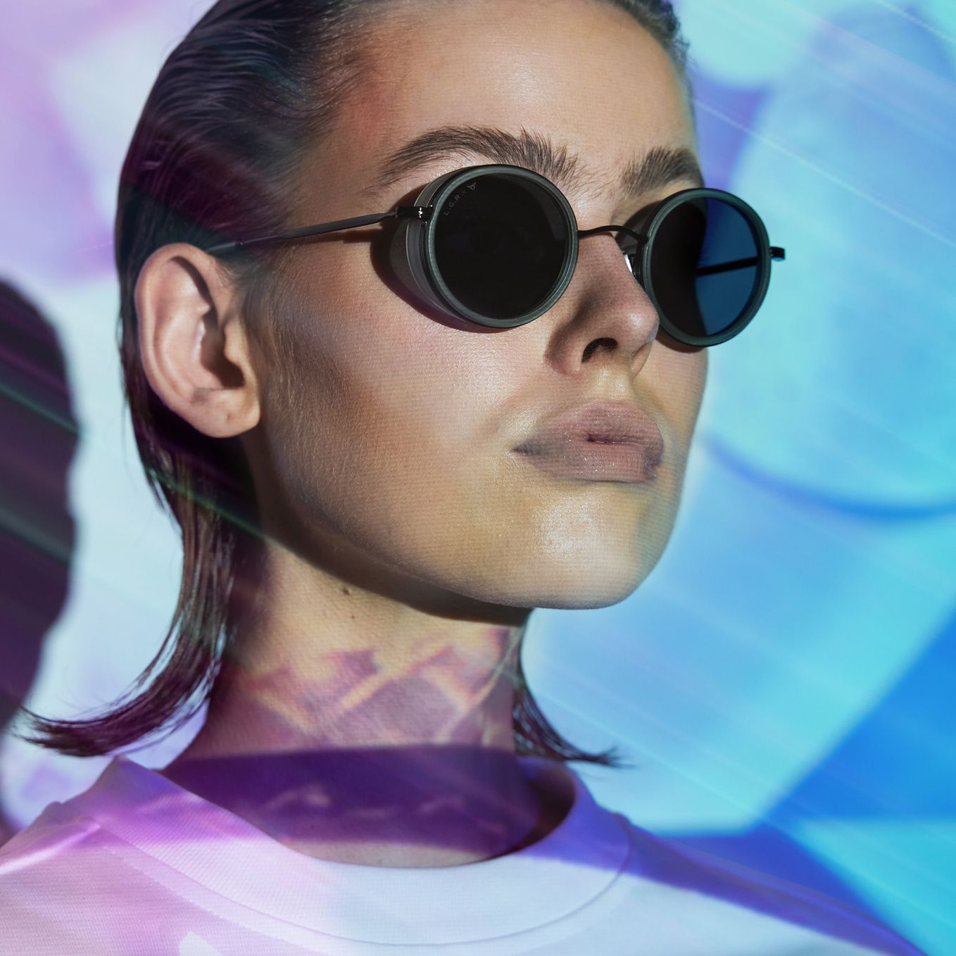 Ženska z gladko počesanimi lasmi in CUPRA sončnimi očali nosi belo majico v osvetljenem okolju z modrimi in rožnatimi svetlobnimi učinki.