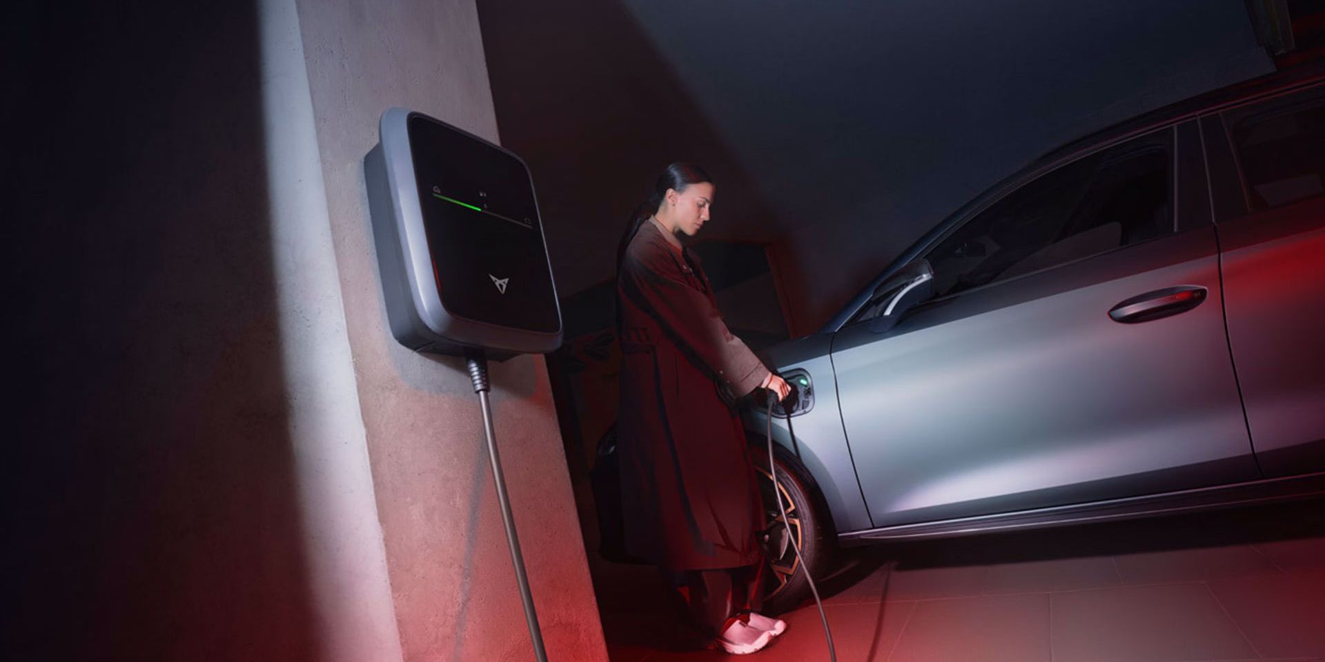 Ženska polni sivi električni avtomobil CUPRA Leon, ki je priključen na polnilno postajo, nameščeno na steni. Okolje je osvetljeno z rdečo in modro svetlobo.