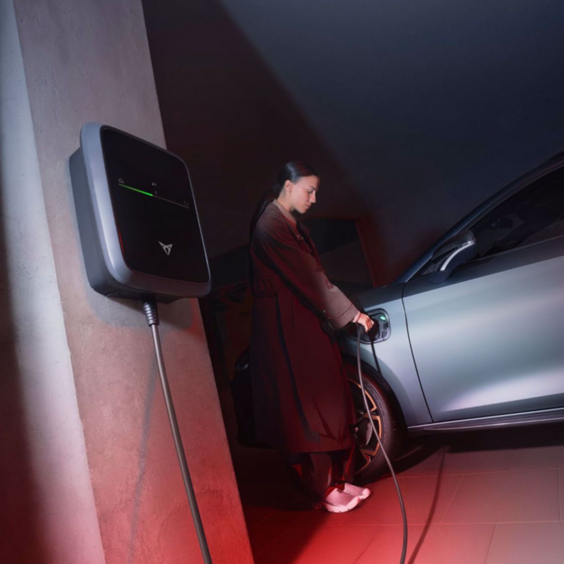 Ženska polni sivi električni avtomobil CUPRA Leon, ki je priključen na polnilno postajo, nameščeno na steni. Okolje je osvetljeno z rdečo in modro svetlobo.