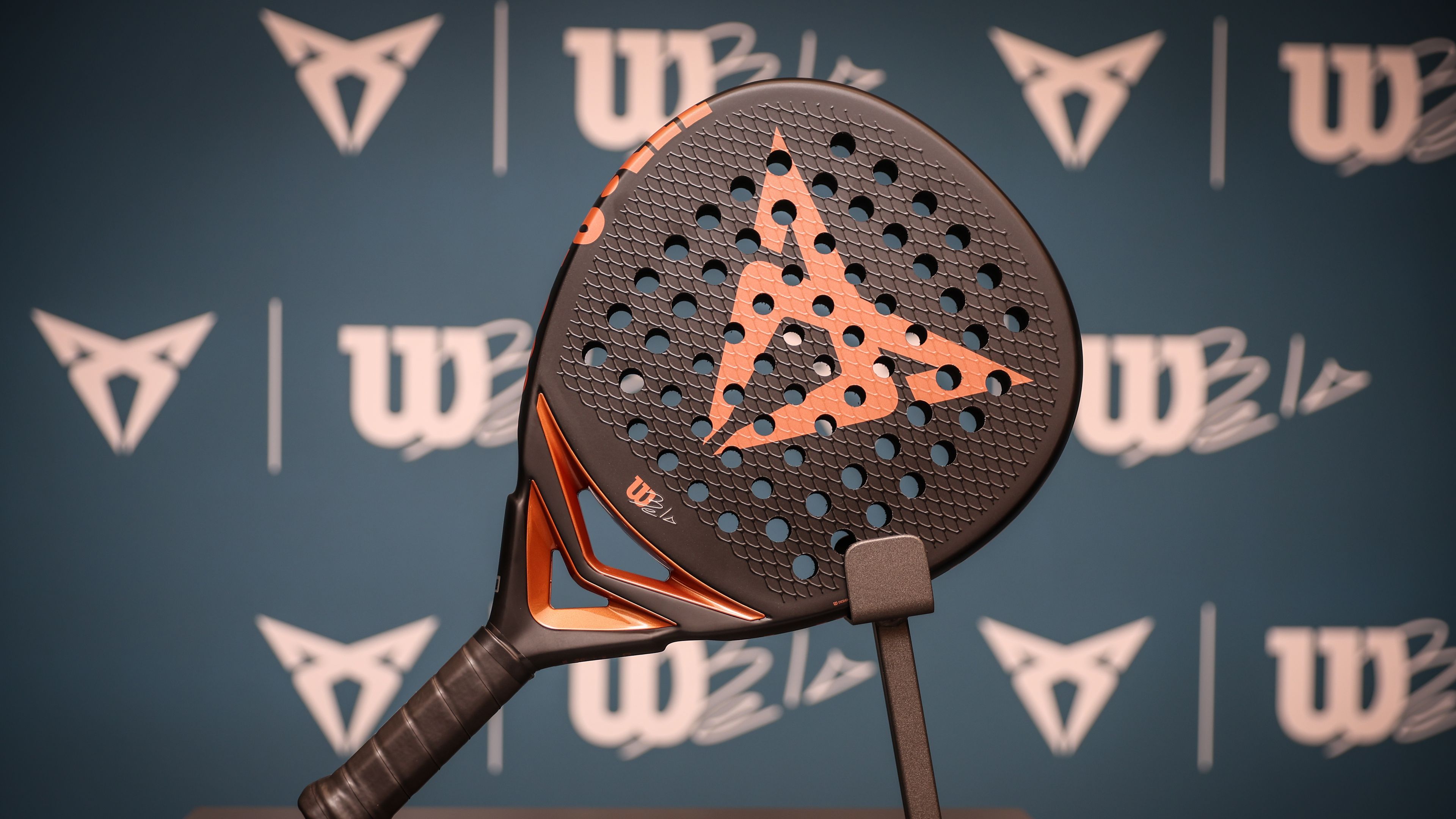 Na stojalu je lopar za padel z logotipom znamke CUPRA v središču. V ozadju so vidni logotipi Wilson, CUPRA in Bela. Lopar je črne barve z oranžnimi detajli.