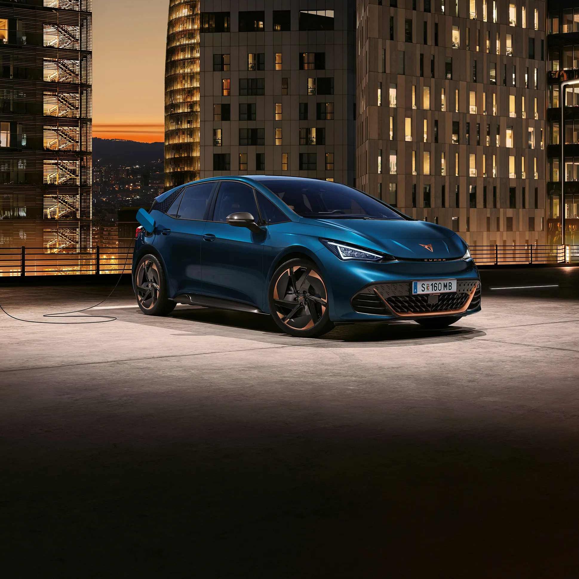 Moderen električni avto CUPRA Born je parkiran na strehi stavbe v urbanem okolju med visokimi zgradbami. V ozadju je vidna osvetljena stolpnica. Avto se polni na polnilni postaji.