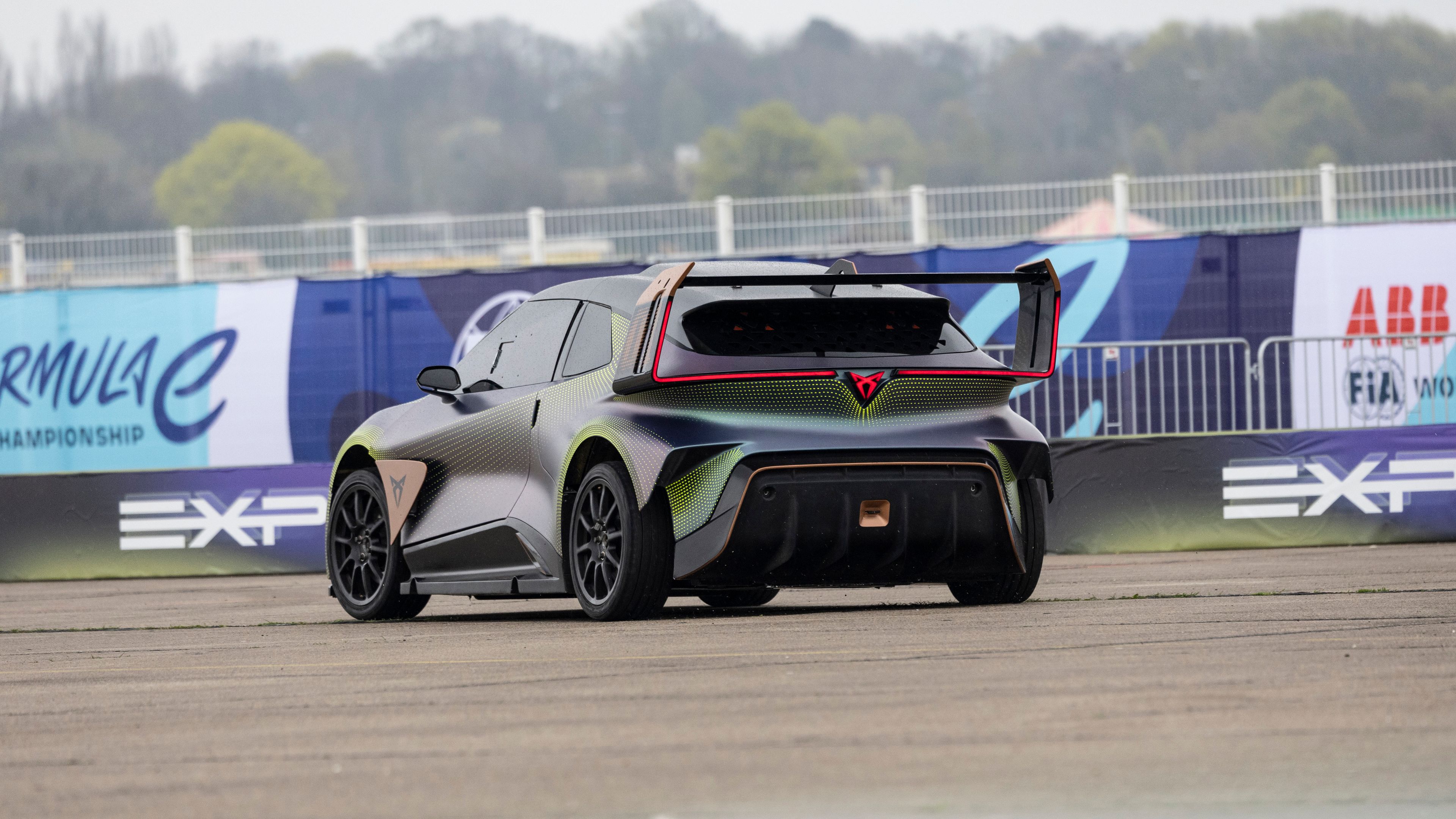 Na sliki je dirkalni avtomobil CUPRA s širokim zadnjim spojlerjem parkiran na asfaltni površini. V ozadju so vidni ograja in panoji dirkaške serije Formula E. V daljavi so vidna drevesa in meglica.