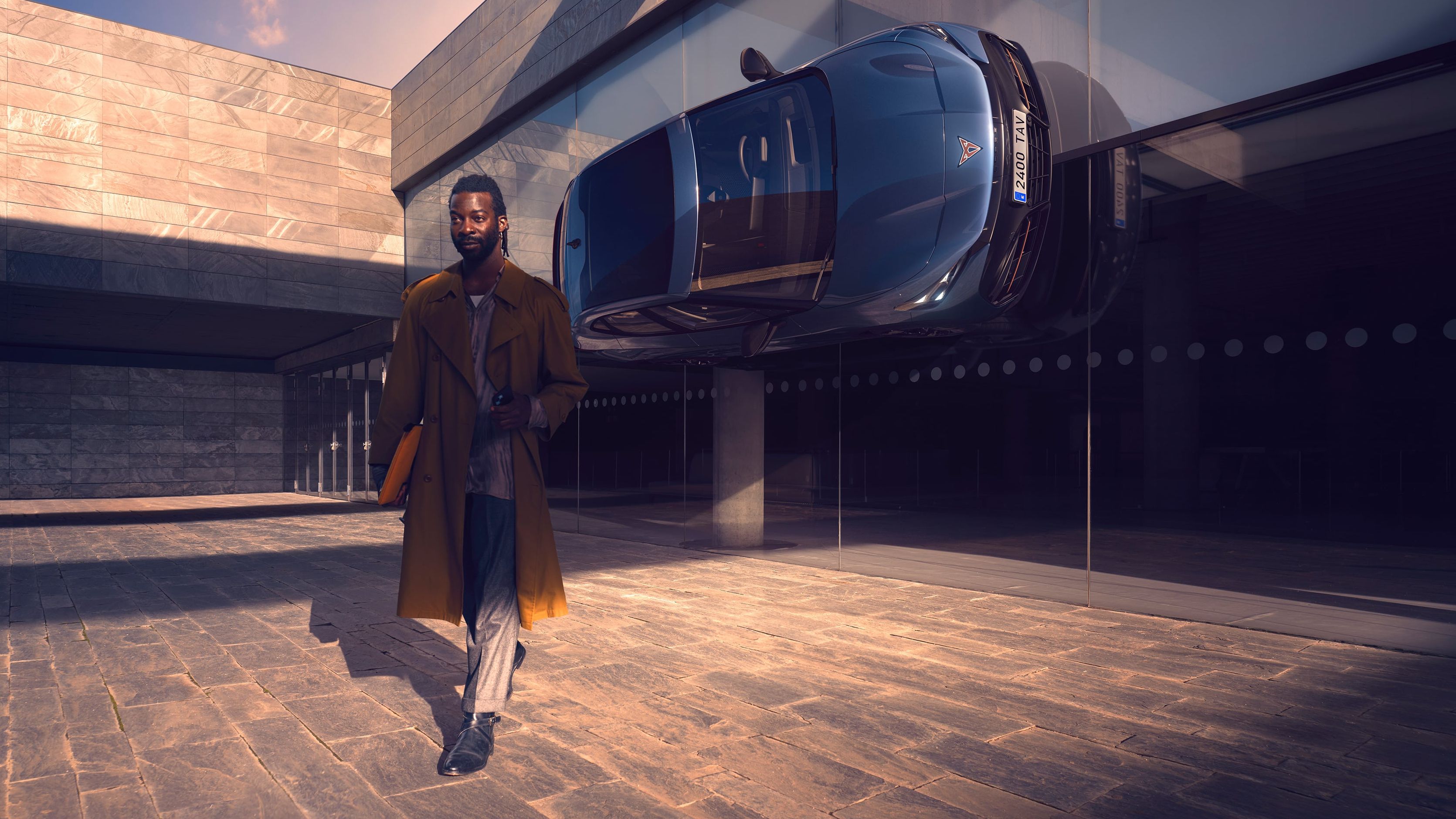 Moški hodi po dvorišču sodobne zgradbe. Ob zgradbi je nameščen modri avtomobil CUPRA Tavascan VZ, ki se zdi, kot da je parkiran navpično na steni. Ozadje sestavljajo steklene površine in kamnite ploščice.