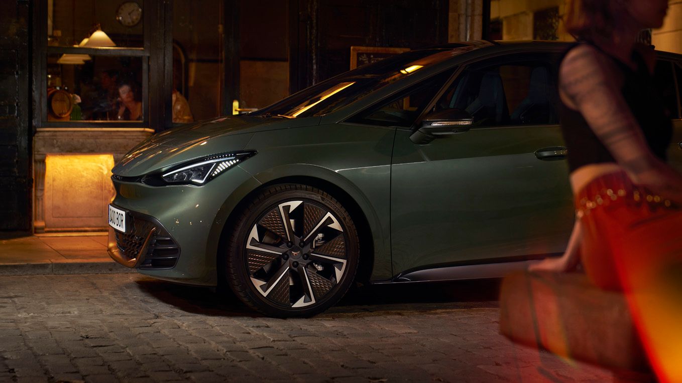 Na sliki je parkiran temno zelen električni avto CUPRA Born pred vhodom v stavbo na tlakovanem pločniku. Zadaj je zamegljena oseba, ki sedi na klopi. Svetloba iz stavbe osvetljuje okolico.