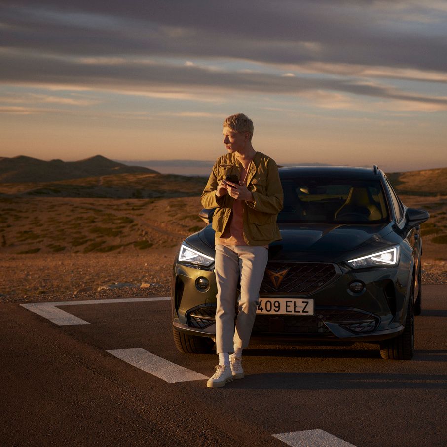 Oseba v svetlih oblačilih stoji pred parkiranim črnim avtomobilom CUPRA Umbrella Formentor, na prazni cesti. Pokrajina v ozadju je hribovita z zemeljskimi toni, pod večernim nebom.