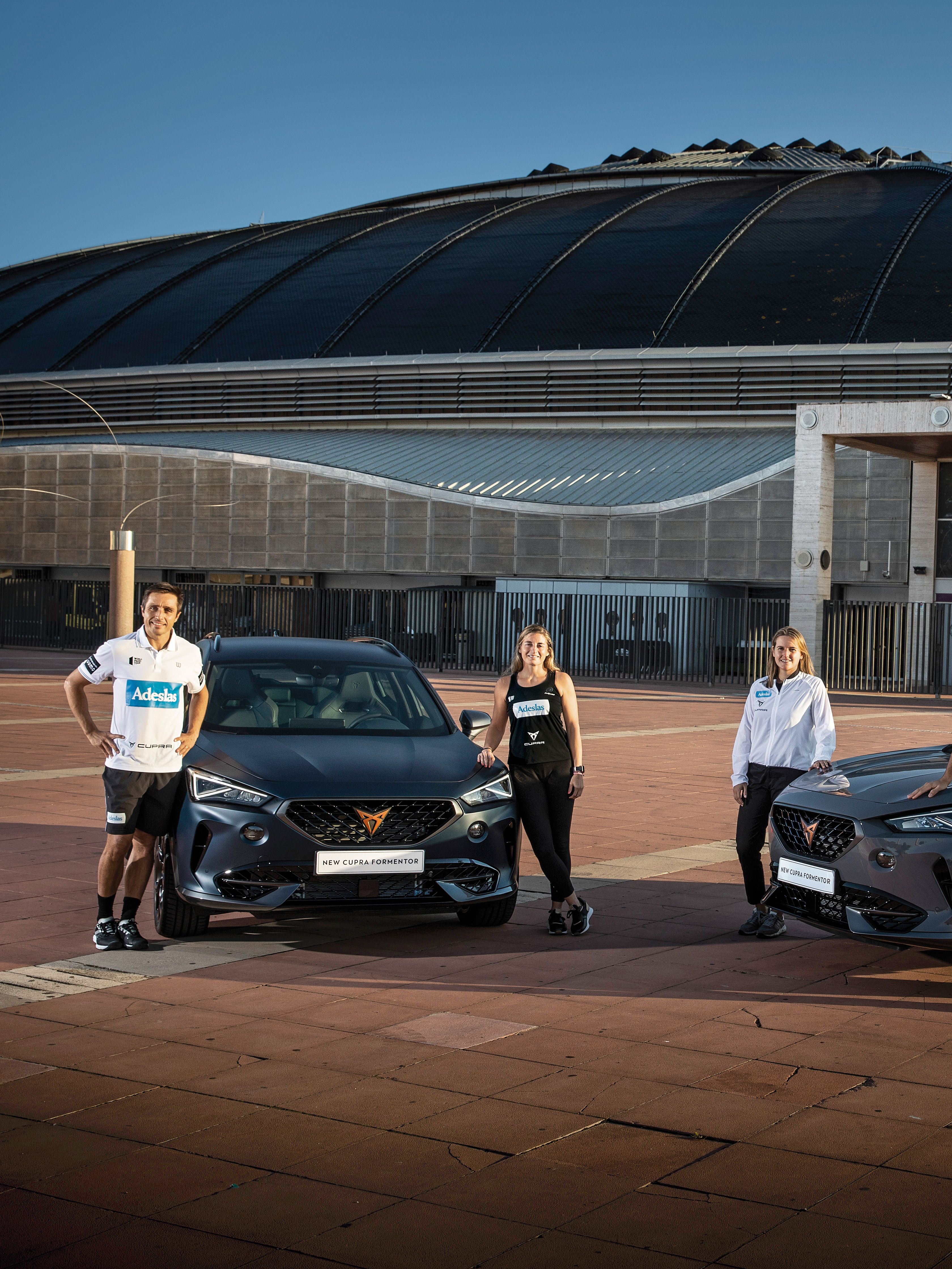 Pet igralcev padel tenisa stoji pred dvema CUPRA avtomobiloma.Za njimi je velika moderna stavba s kupolsko streho.