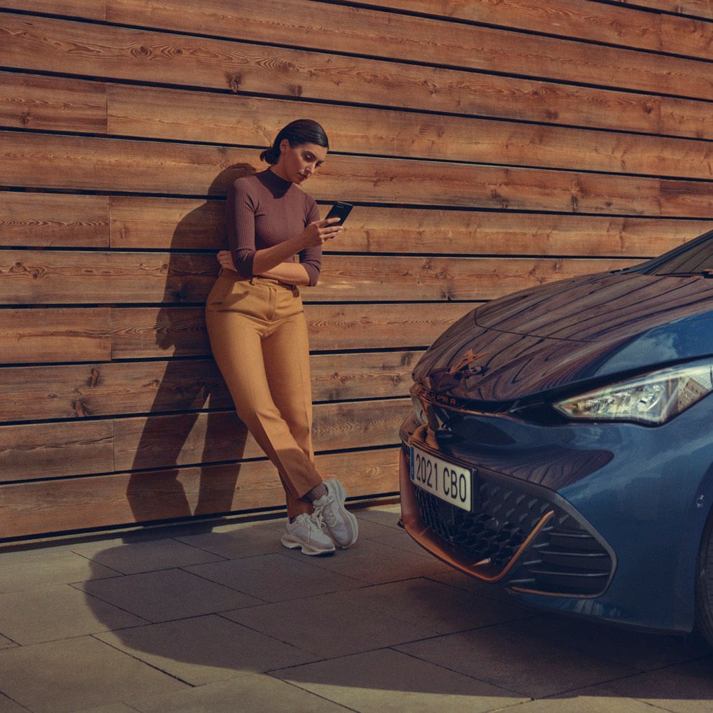 Ženska, stoji poleg avtomobila CUPRA Born, modre barve. Naslonjena  je na leseno steno in gleda v telefon. 