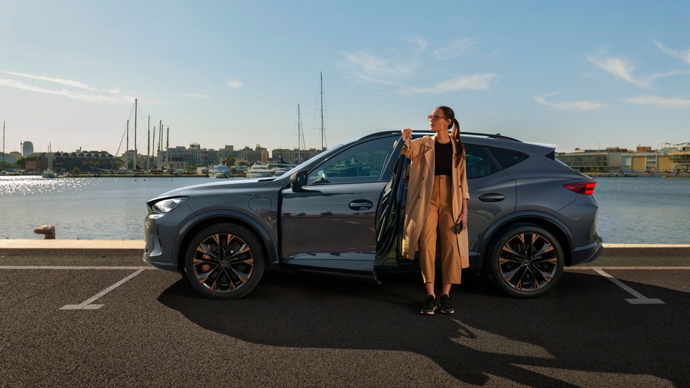 Ženska stoji poleg parkiranega sivega avtomobila CUPRA Formentor ob vodi in drži se za odprta vrata. V ozadju so jadrnice na pristanišču in zgradbe. 