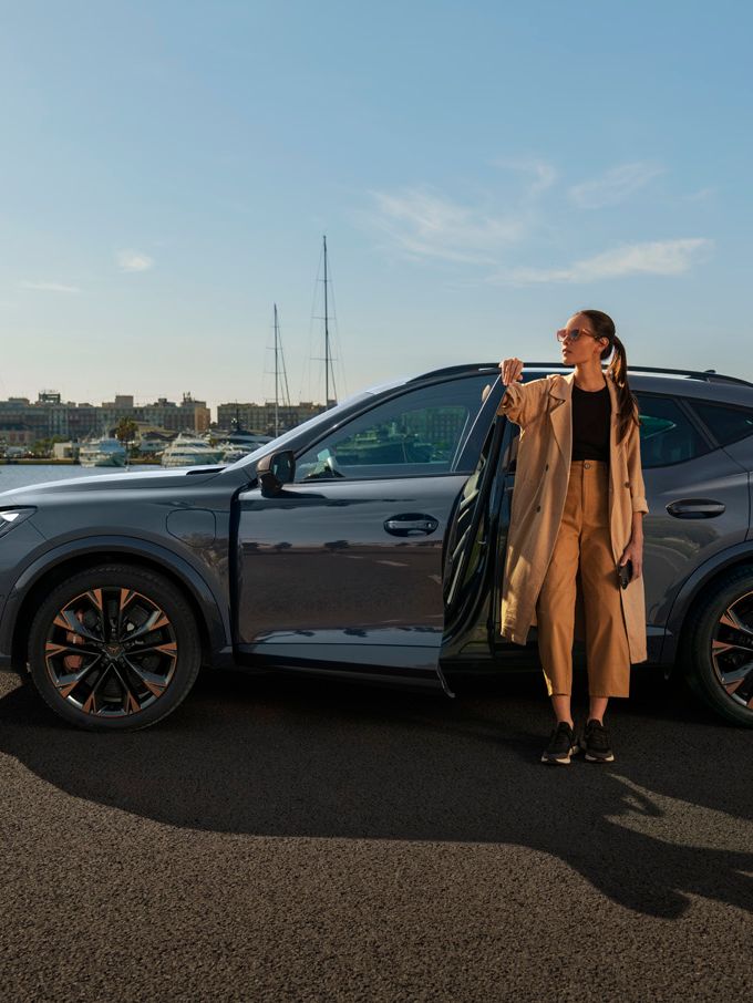 Ženska stoji poleg parkiranega sivega avtomobila CUPRA Formentor ob vodi in drži se za odprta vrata. V ozadju so jadrnice na pristanišču in zgradbe. 