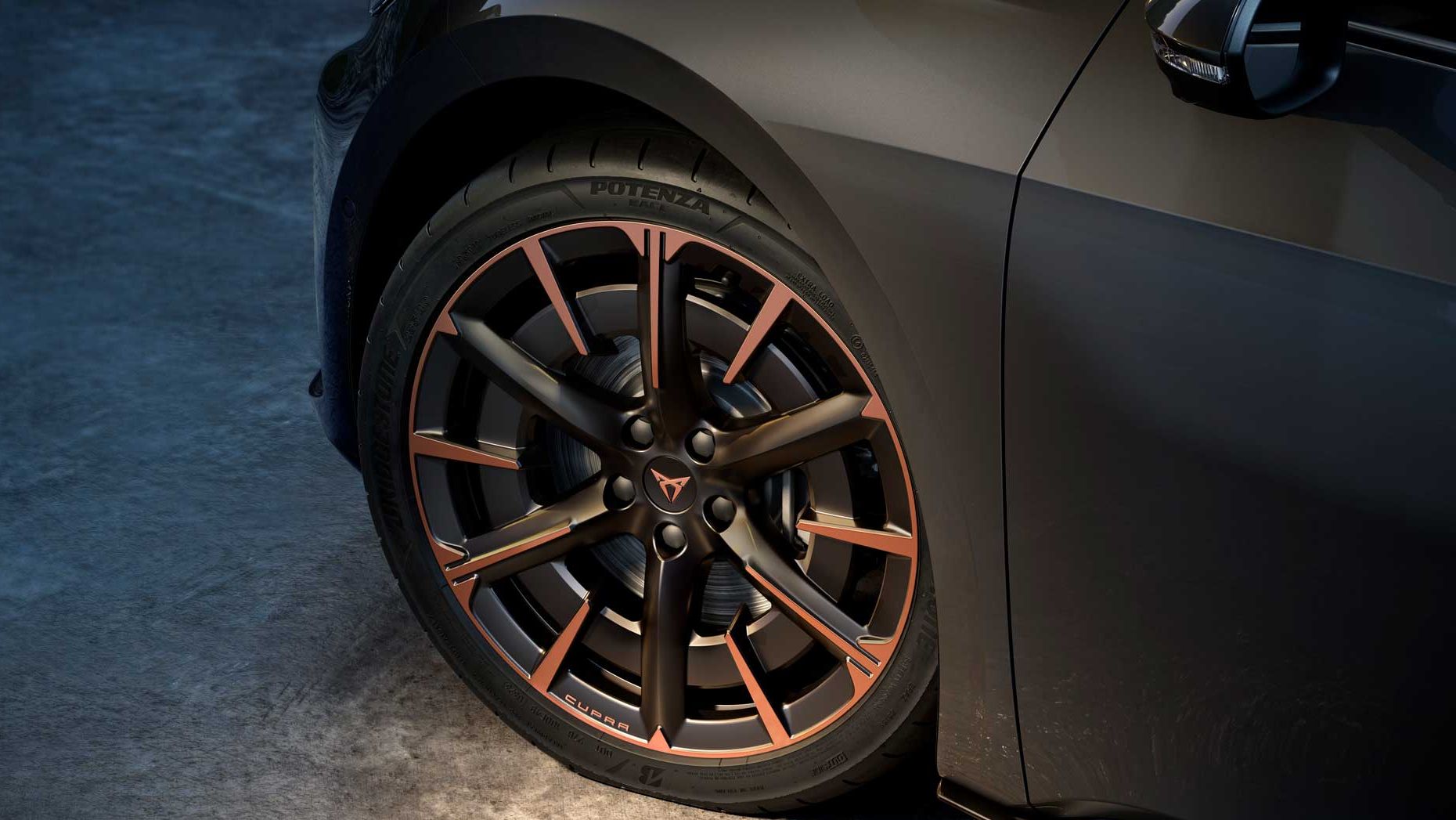 Detaljen sprednjega kolesa CUPRA Born z velikimi športnimi platišči v mat črni barvi z bakrenimi poudarki. Avto je temno sive barve, osvetljen od spodaj, kar poudari obliko platišča in pnevmatike. 