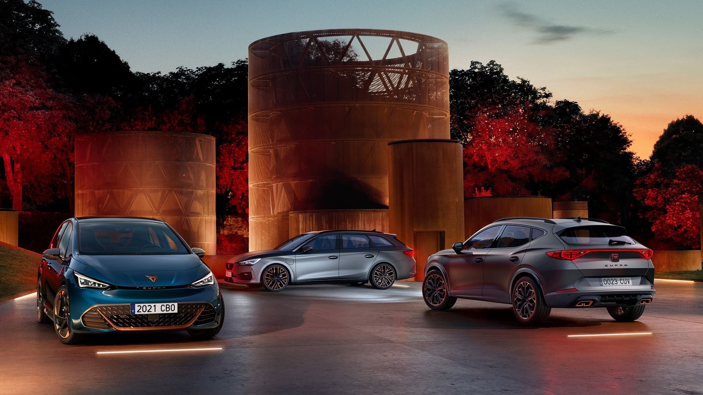 Tri sodobni avtomobili - CUPRA BORN, CUPRA LEON SP, CUPRA FORMENTOR so parkirani na dvorišču ob velikih kovinskih skulpturah v času sončnega zahoda. 
