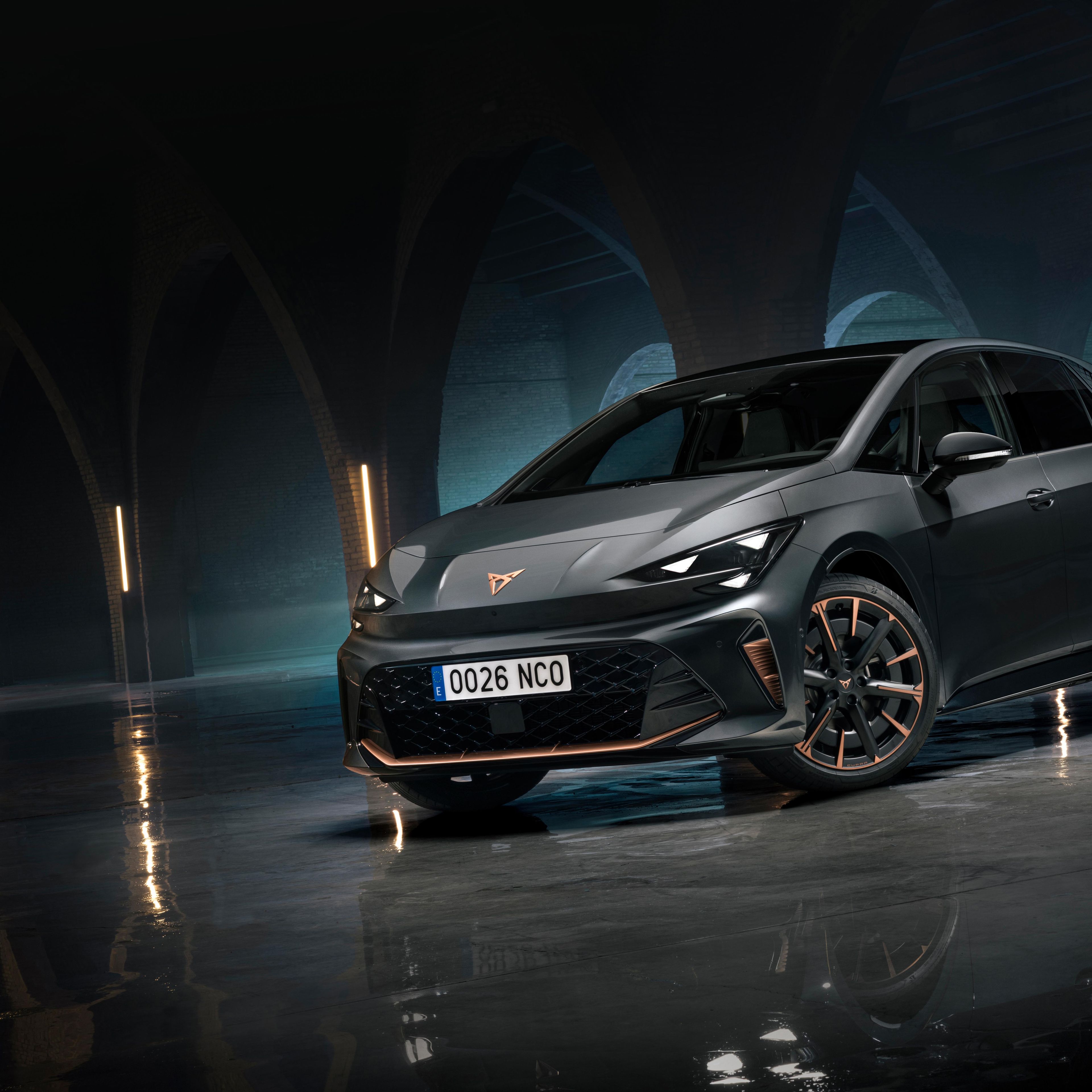 CUPRA Born stoji v temnem, arhitekturno izrazitem prostoru z oboki in refleksijo na mokrih tleh. Dramatična osvetlitev poudarja športen dizajn in električni značaj vozila.