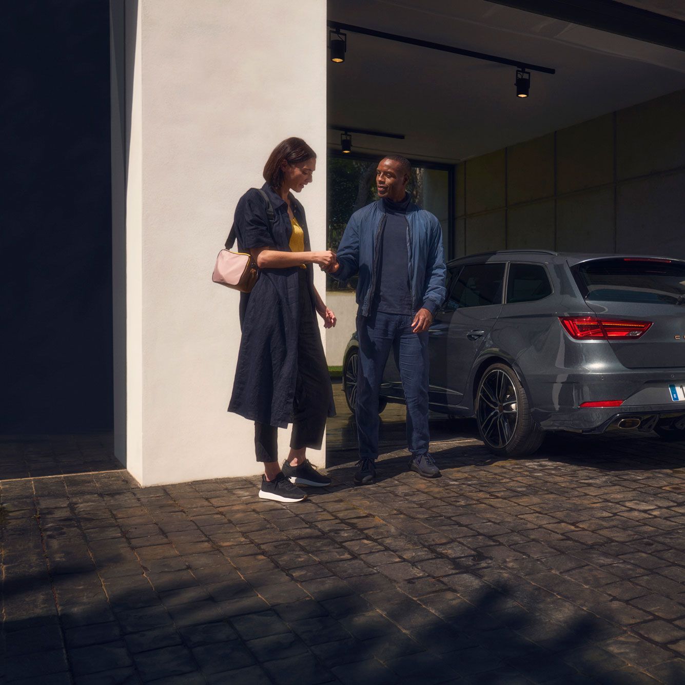 Ženska in moški stojita pred sodobno hišo, na dovozu pa je parkiran temno sivi avto SEAT Leon CUPRA.