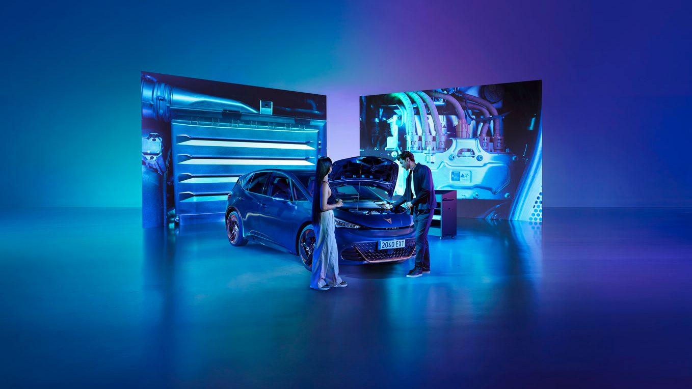 Ženska in moški stojita v studiu ob modrem avtomobilu CUPRA Born z odprtim pokrovom motorja v svetlo modri osvetlitvi. Za avtomobilom so veliki zasloni, ki prikazujejo dele motorja.