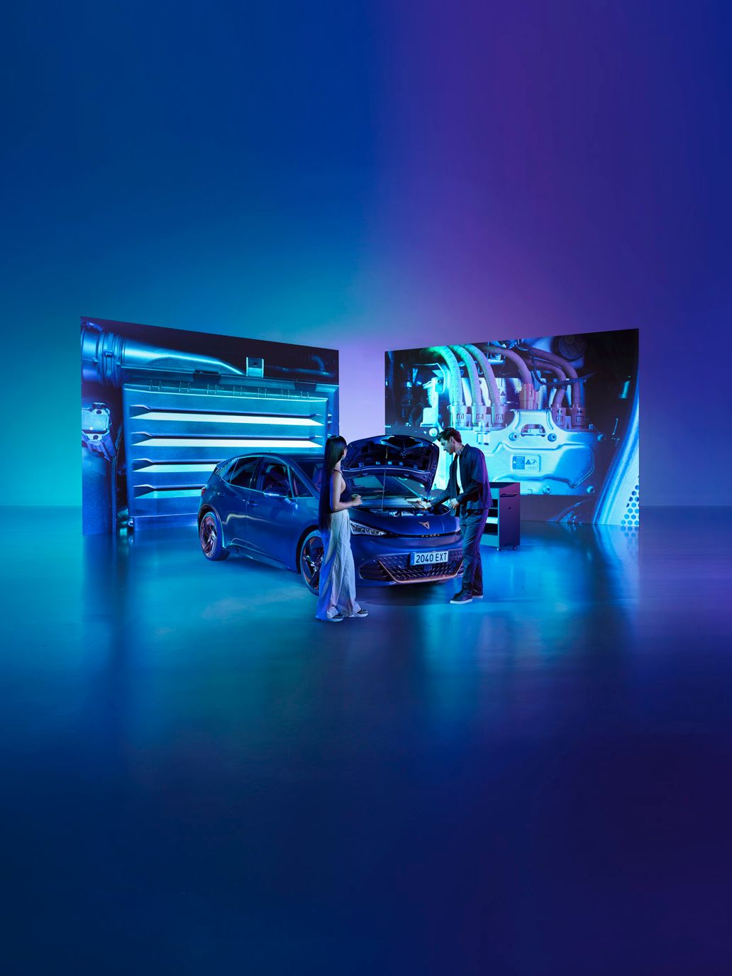 Ženska in moški stojita v studiu ob modrem avtomobilu CUPRA Born z odprtim pokrovom motorja v svetlo modri osvetlitvi. Za avtomobilom so veliki zasloni, ki prikazujejo dele motorja.