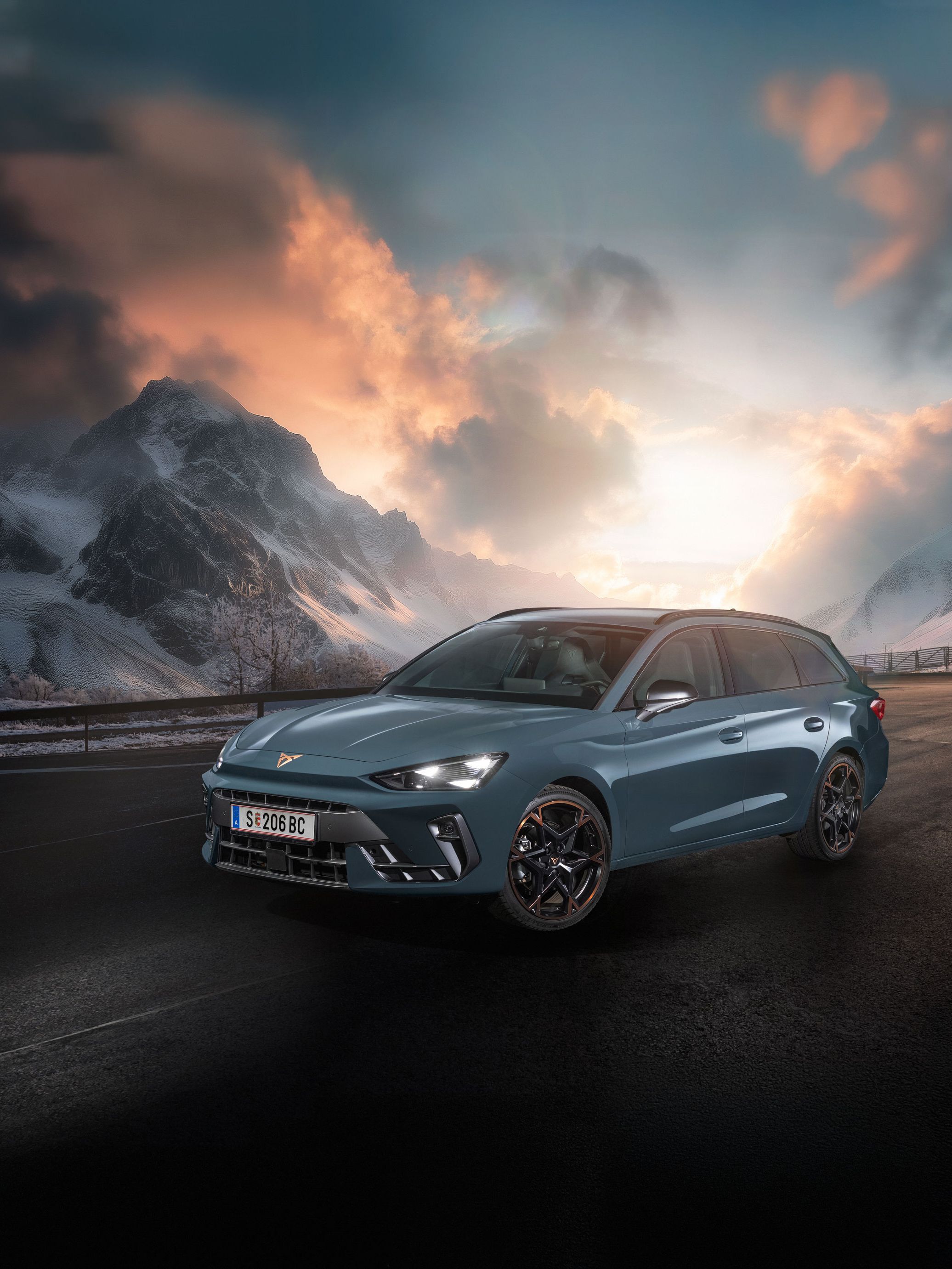 Ein CUPRA Leon Kombi parkt vor einer malerischen Berglandschaft bei Sonnenuntergang. Im Hintergrund sind verschneite Gipfel und dramatische Wolken zu sehen. Der Asphalt ist dunkel und feucht.