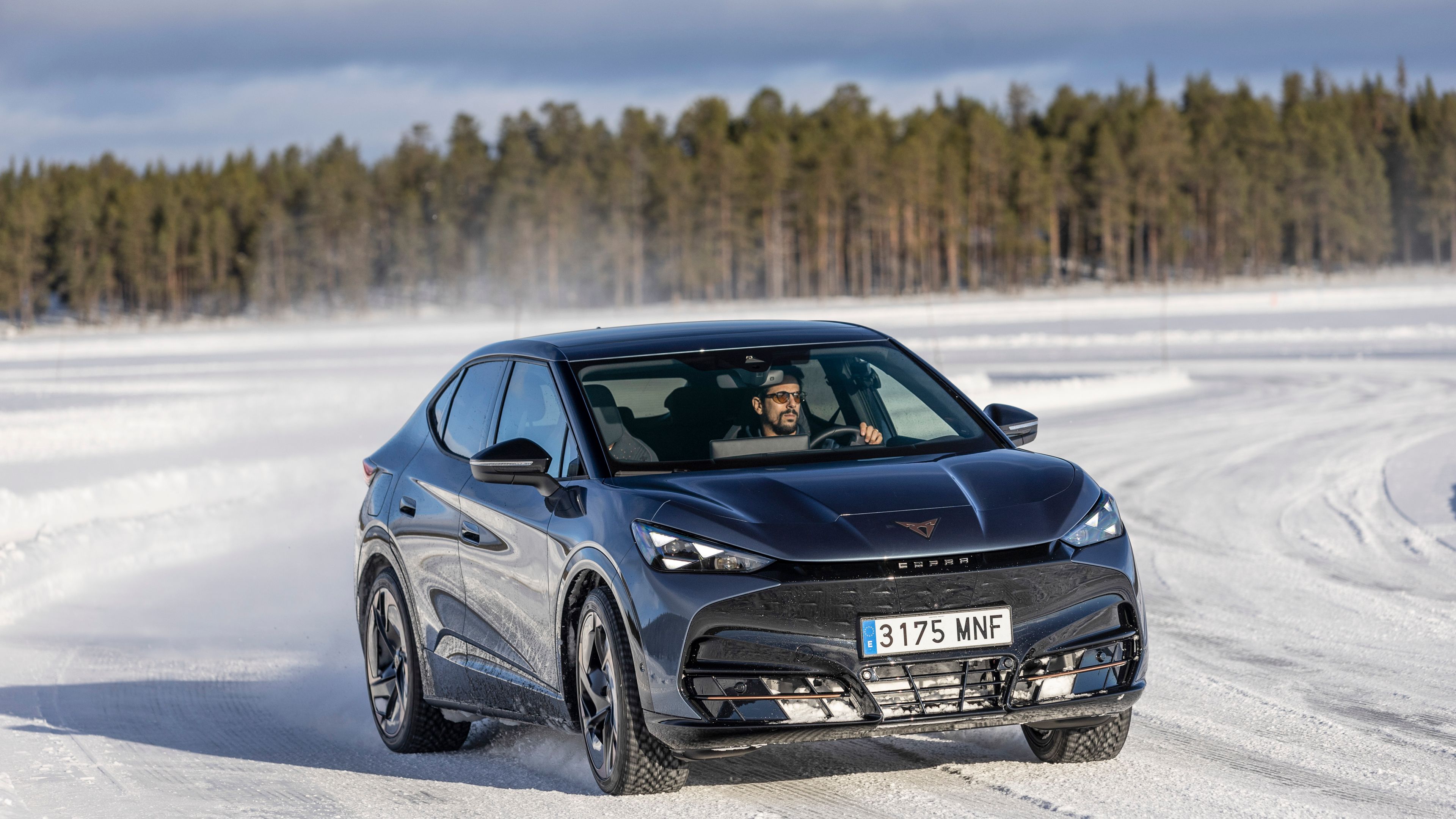 Lucas di Grassi vozi CUPRA Tavascan po zasneženi cesti, medtem ko padajo zimski sončni žarki. V ozadju je gozd dreves. Avto dviguje sneg, kar kaže na hitrost vožnje.