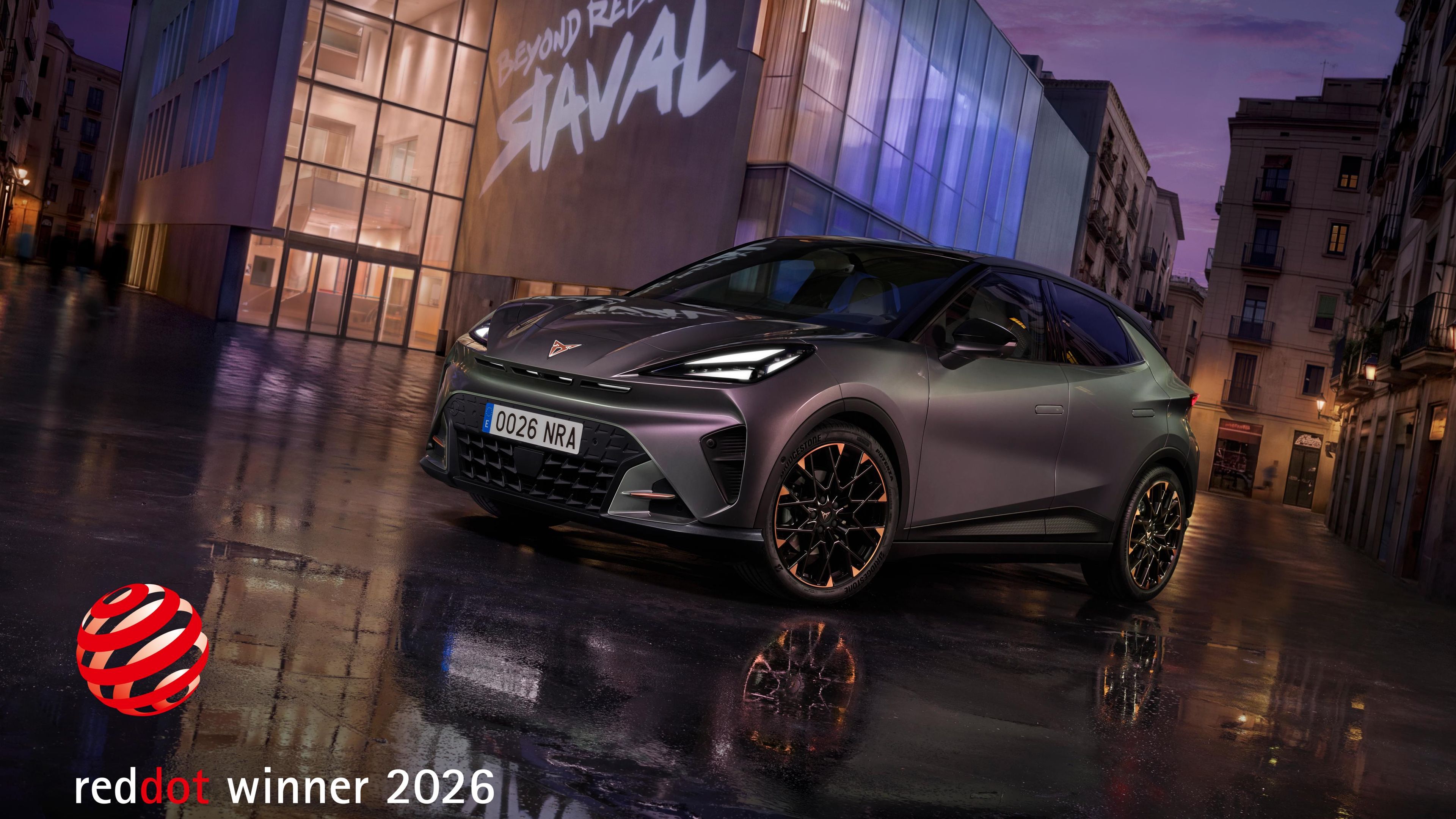CUPRA Raval stoji na mokri mestni ulici pred sodobno stavbo z napisom »Raval« v večerni svetlobi. Oznaka »Red Dot Winner 2026« poudarja nagrajeni dizajn vozila.
