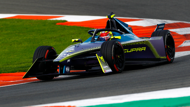 Dirkalnik formula E zavija okoli ovinka na dirkališču. Avto je moder z rumenimi in zelenimi poudarki ter napisom "CUPRA" na strani. Voznik nosi čelado in dirka po asfaltni stezi ob zeleni trati.