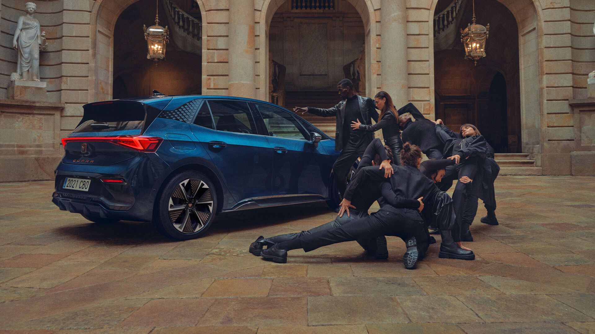 Skupina elegantno oblečenih ljudi izvaja plesne poteze poleg Cupra Borna v notranjem dvorišču zgodovinske zgradbe.