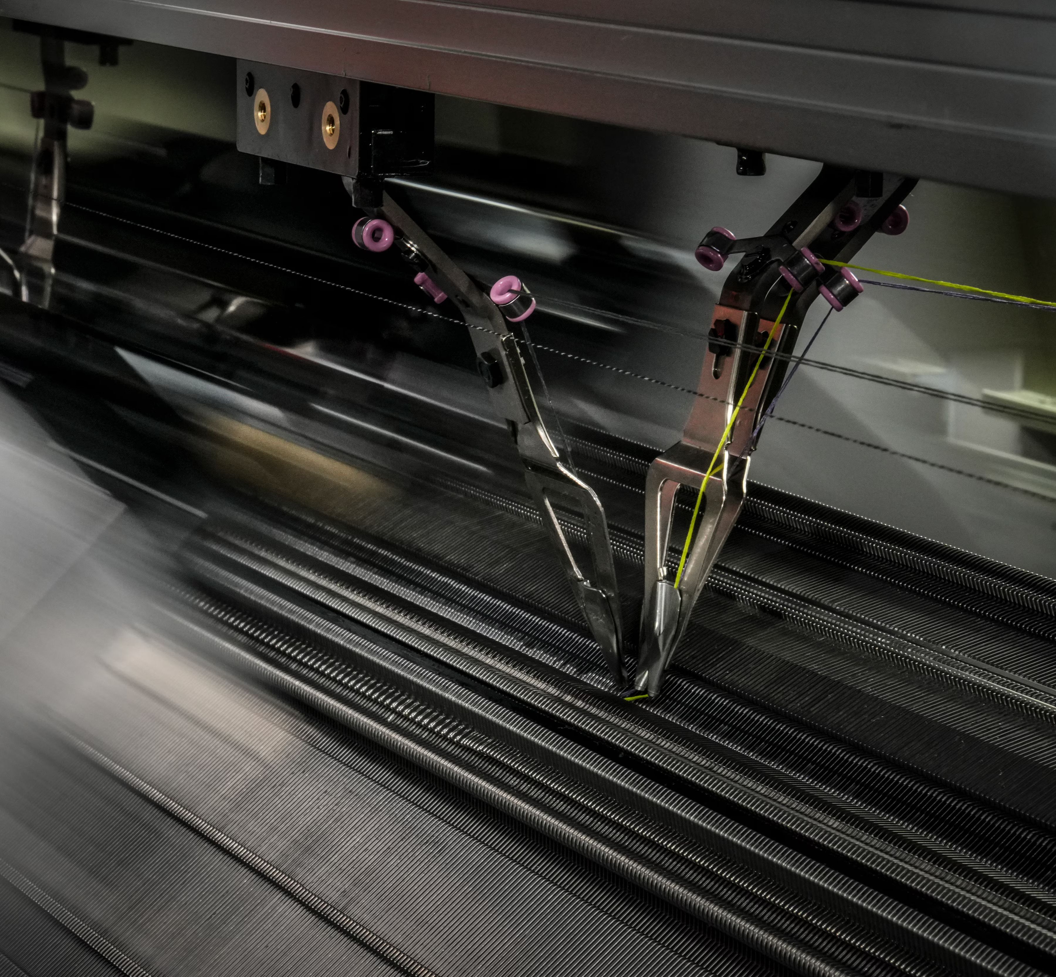 Industrijski 3D stroj v gibanju tiska oziroma plete tehnični tekstilni material. Vidni so mehanski elementi in niti, ki prikazujejo natančen proces avtomatizirane izdelave.