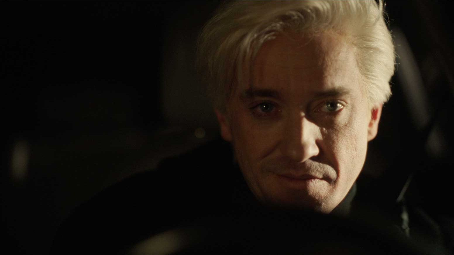 Tom Felton sedi v zatemnjenem prostoru vozila in gleda proti kameri. Mehka svetloba poudarja dramatično in skrivnostno vzdušje prizora.
