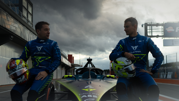 Dva dirkača v dirkalnih oblekah sedita na dirkalnem avtomobilu na dirkališču. Oba držita čeladi v rokah. V ozadju je videti stavbo ob dirkališču in oblačno nebo.