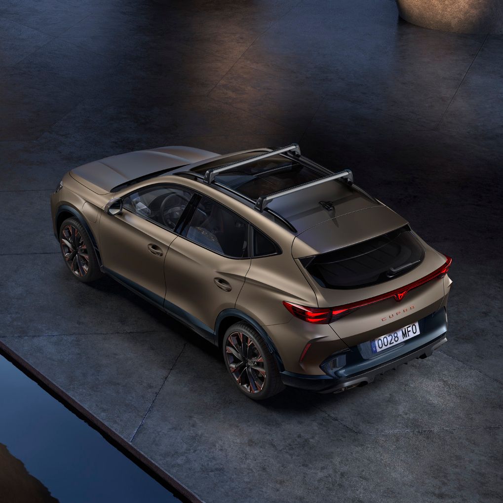 Avtomobil CUPRA Formentor PA stoji na sodobnem parkirišču. Vozilo je v kovinsko rjavi barvi z nosilcem na strehi. Ima izrazita zadnja svetila in športen dizajn.