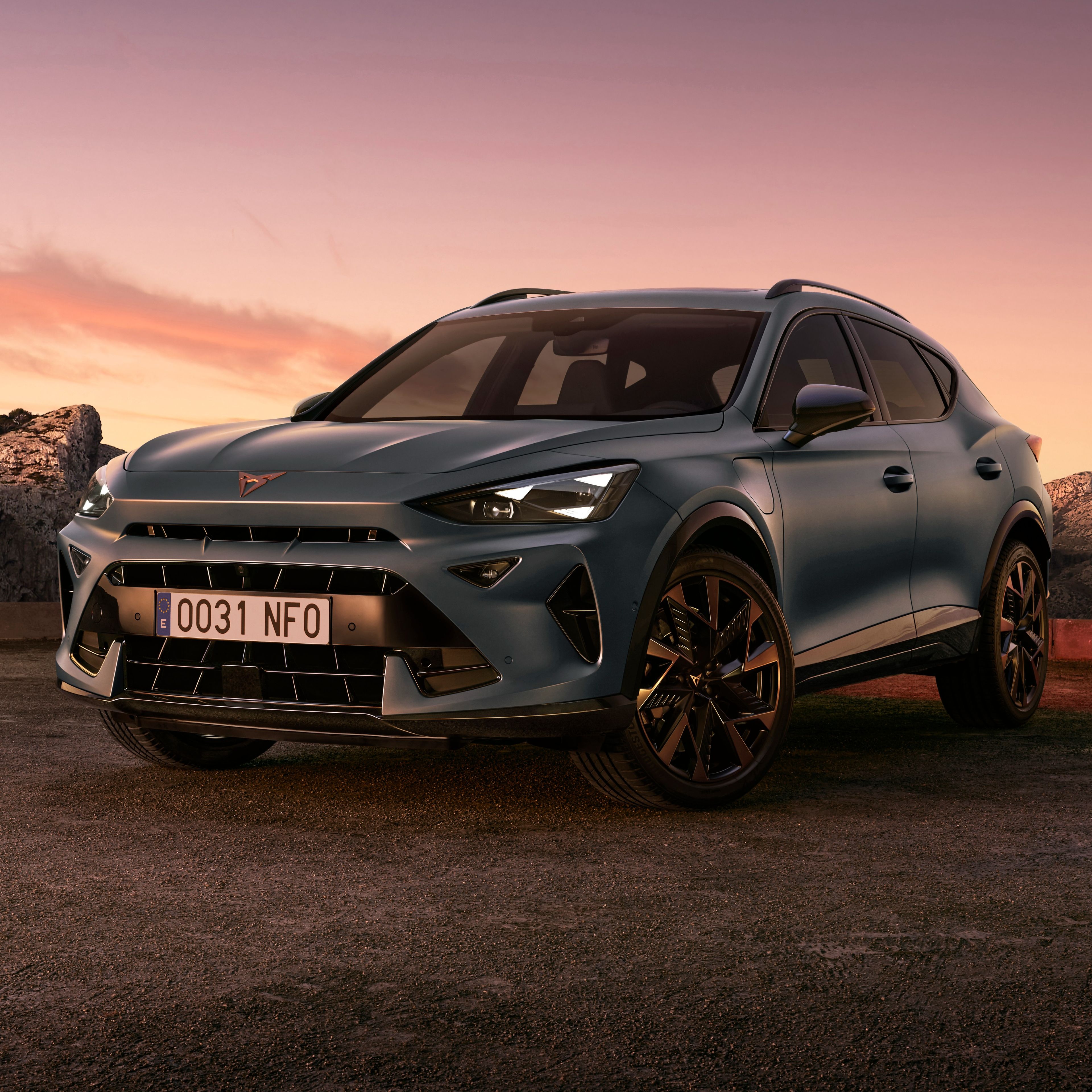 CUPRA Formentor stoji na prostornem parkirišču ob skalah med sončnim vzhodom ali zahodom. Nebo je obarvano v tople odtenke vijolične in oranžne barve.