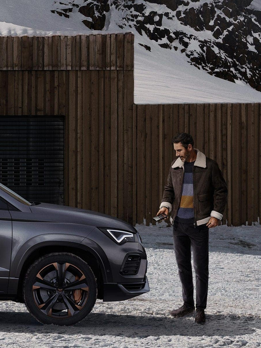 Moški stoji poleg temno sivega vozila CUPRA Ateca, parkiranega pred leseno zgradbo v zasneženem okolju na gori.